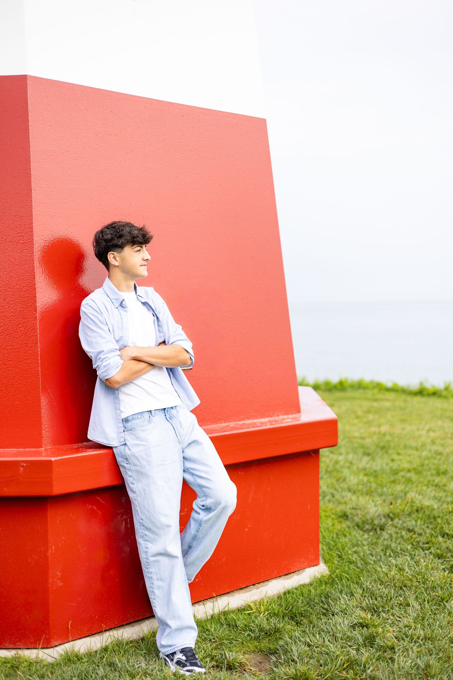 Young man with curly hair gazing thoughtfully outdoors near green trees by water.