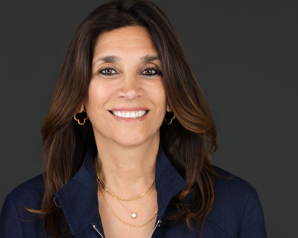 Professional headshot of a smiling woman with long brown hair wearing a navy top and gold jewelry.