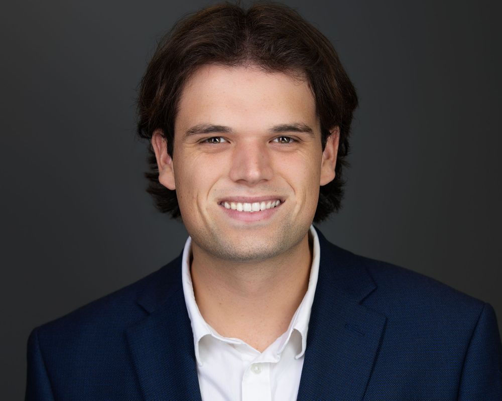 Young professional man smiling in navy blazer and white shirt against dark background, corporate headshot.