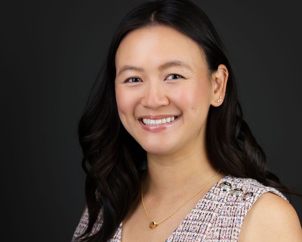 Professional headshot of a smiling young Asian woman with dark hair, wearing a tweed top and gold necklace.