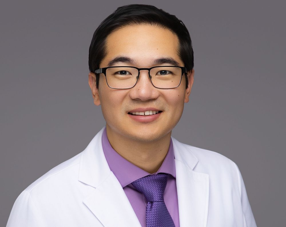 Professional male doctor in white lab coat and purple tie smiling confidently for a medical practice headshot.
