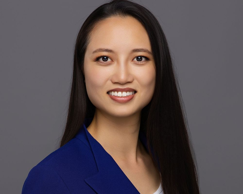 Professional headshot of a smiling young woman in a blue blazer against a gray background.