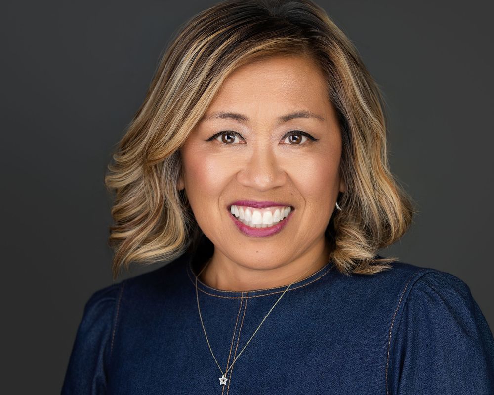 Professional headshot of a smiling woman with highlighted hair wearing a denim top and delicate necklace against a dark background.