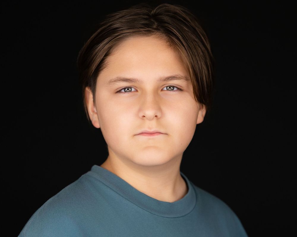 Young person with brown hair wearing a teal shirt, posing for a professional headshot on dark background.
