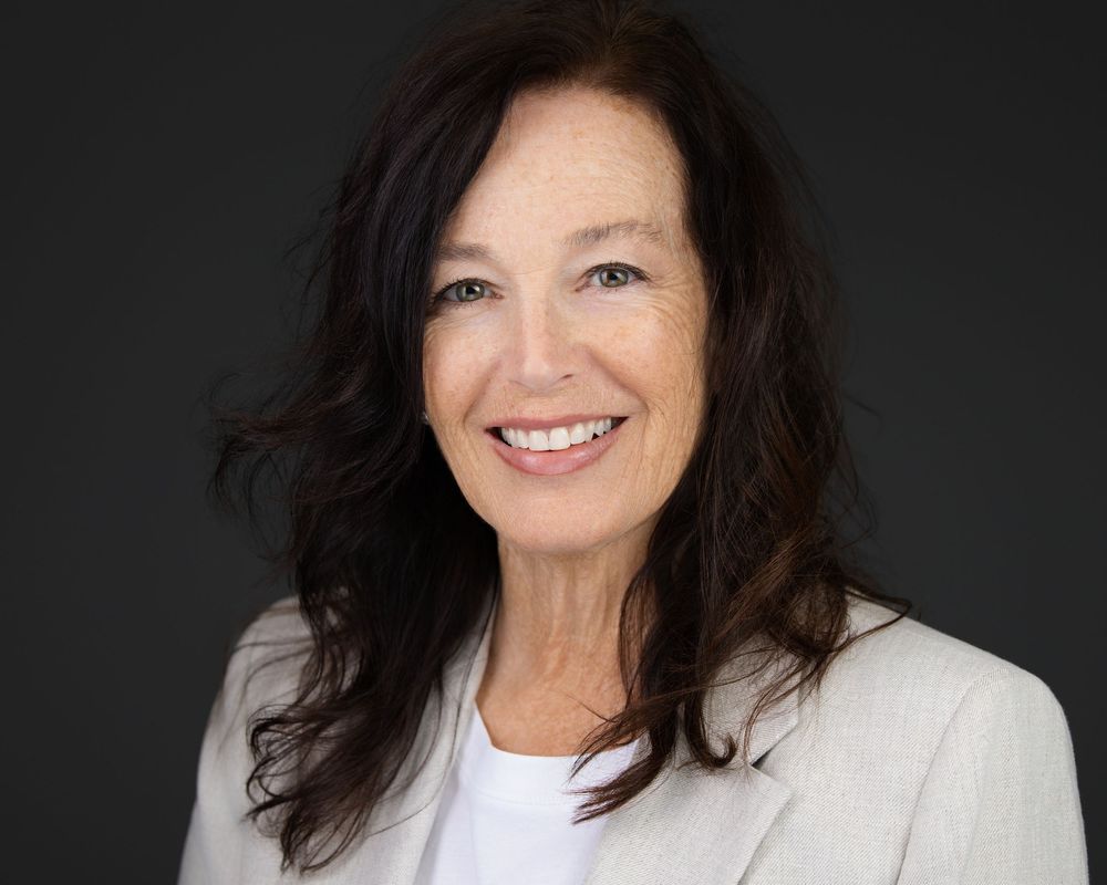 Professional headshot of a smiling middle-aged woman with dark hair wearing a white blazer against a dark background.