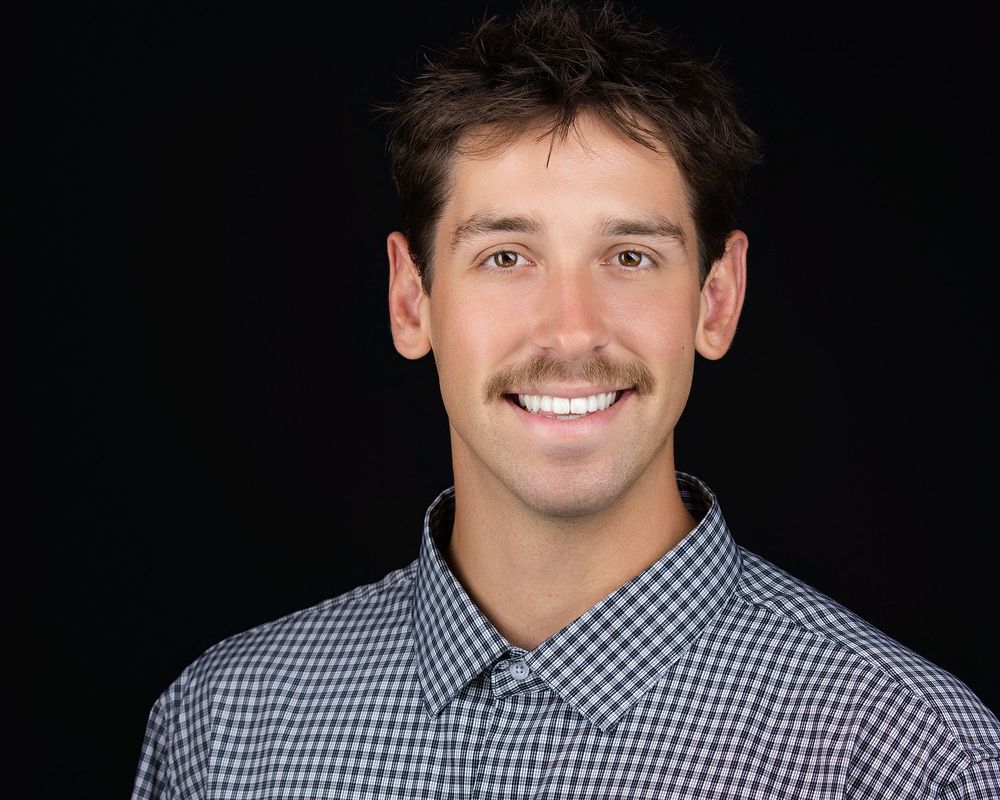 Smiling young man with mustache wearing a blue plaid shirt against a dark background.