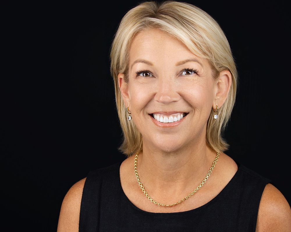 Professional headshot of a smiling blonde woman wearing a black top and gold necklace against a dark background.