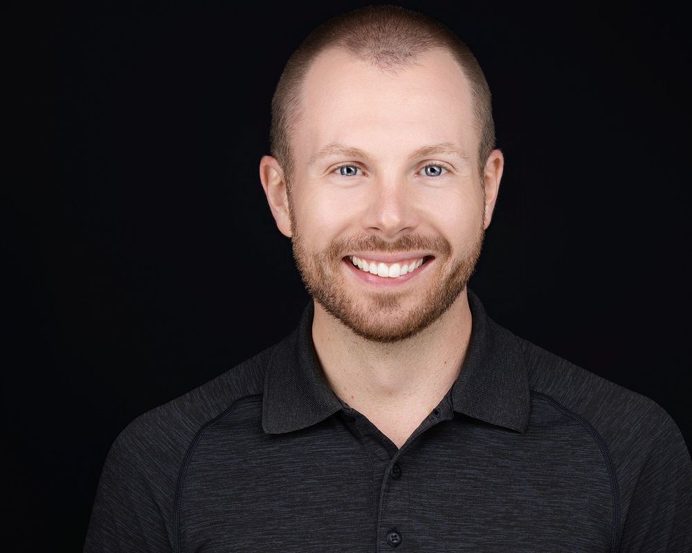 Professional headshot of a smiling man with short hair and beard wearing a dark polo shirt against black background.