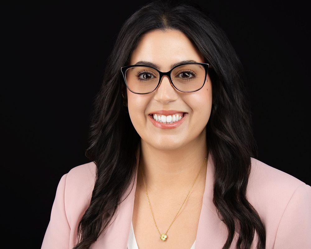 Professional headshot of a smiling woman with dark hair and glasses wearing a pink blazer.