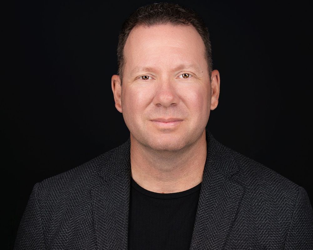 Professional headshot of a confident middle-aged man in a dark blazer and black shirt against a black background.