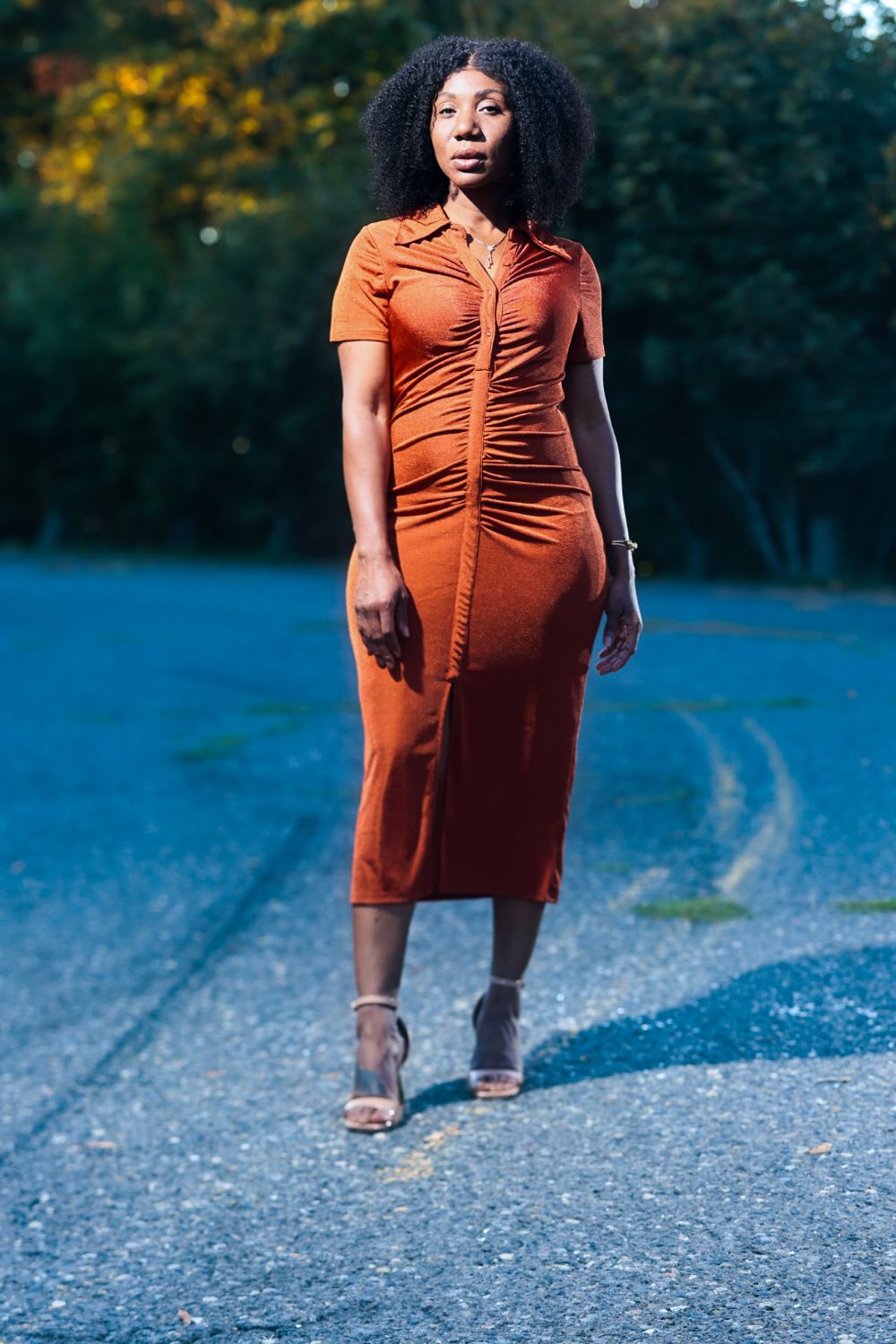 Woman in rust orange midi dress standing on gravel road with trees in background at dusk.