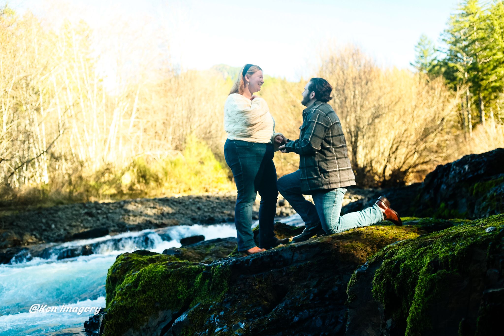 Man proposing to woman on moss-covered rocks beside a flowing river surrounded by autumn trees.