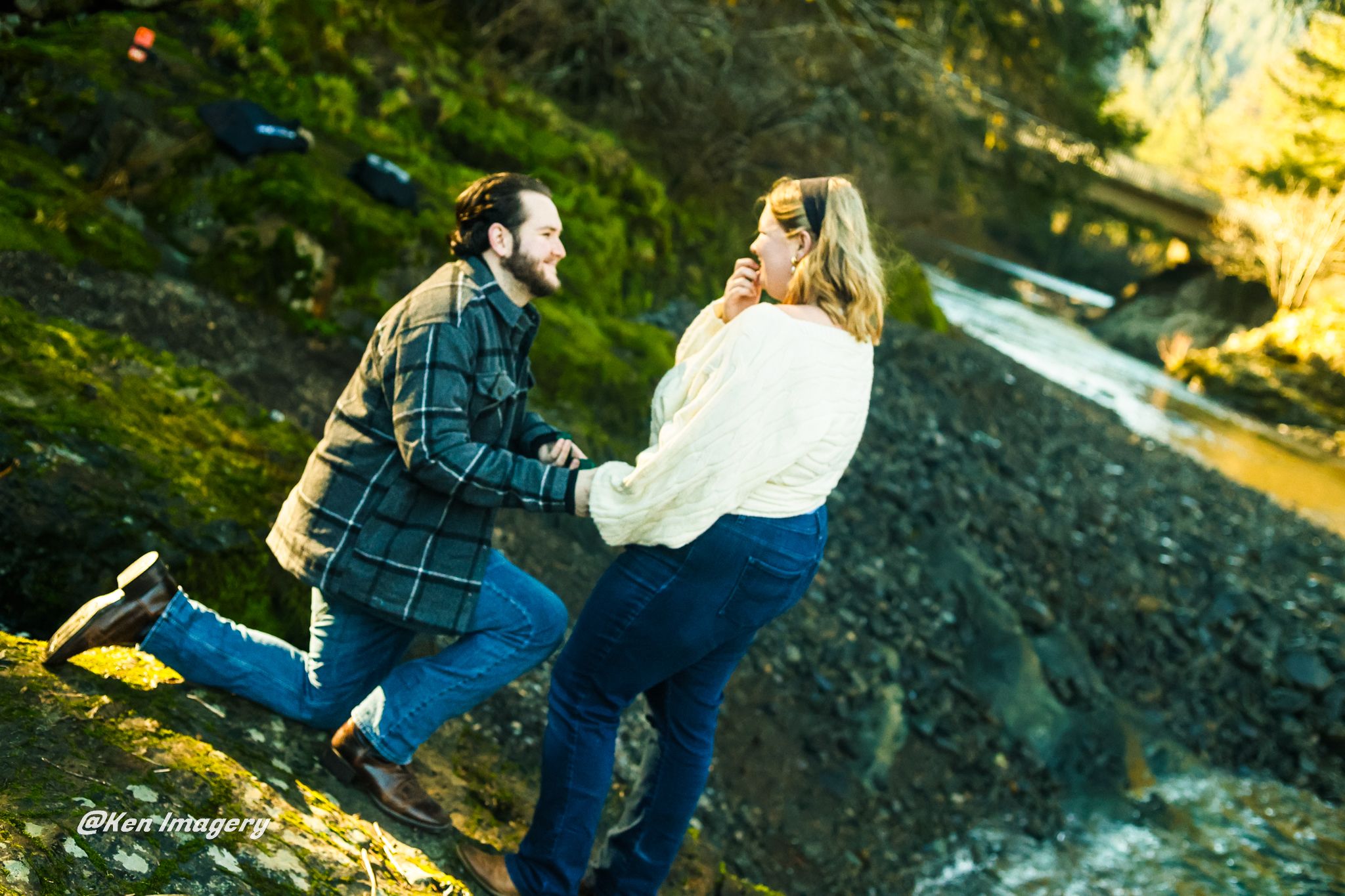 Couple holding hands and dancing outdoors near a rocky stream surrounded by lush green moss and trees.