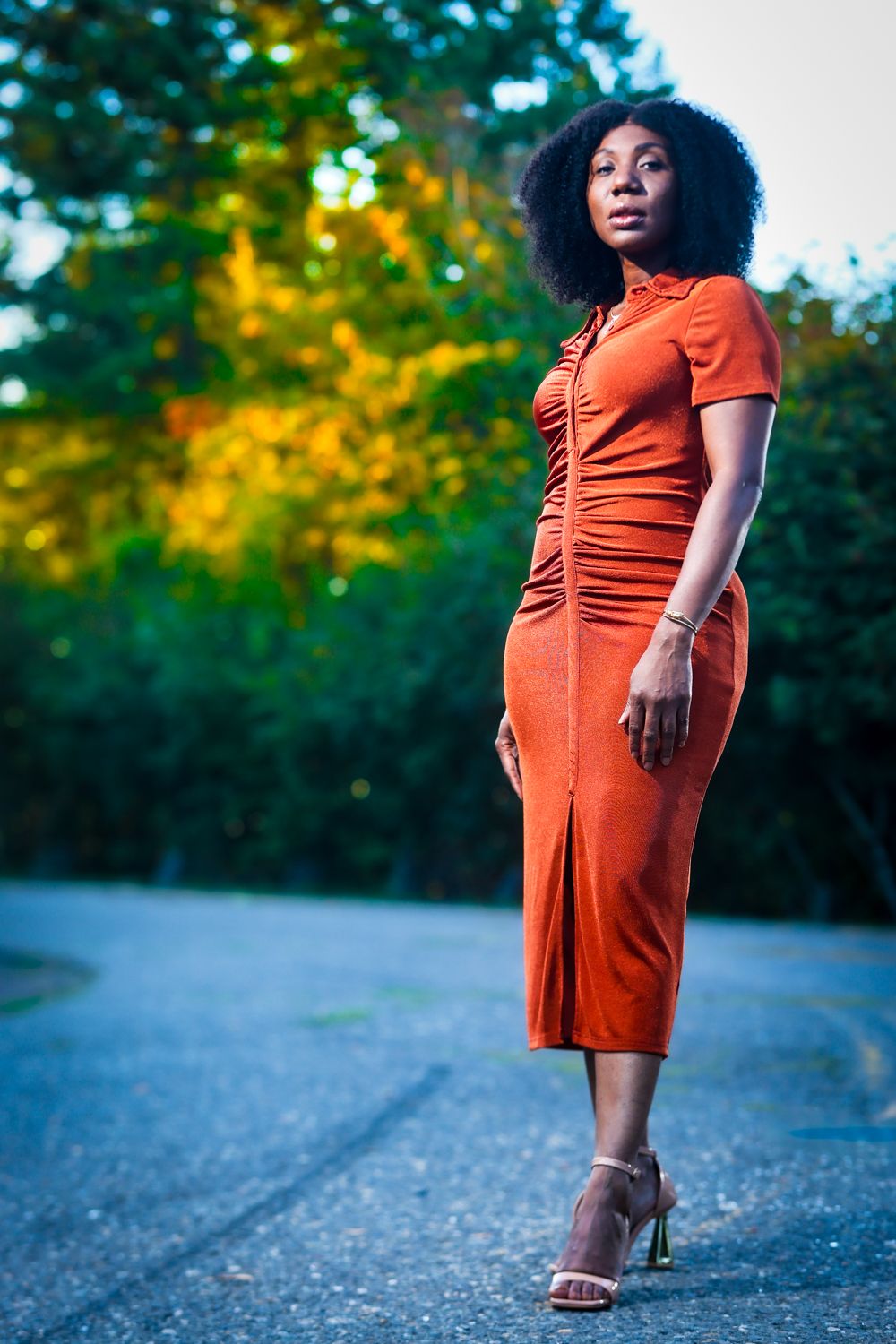A Black woman in a rust-orange midi dress and heels poses confidently on a road with colorful autumn trees behind her.