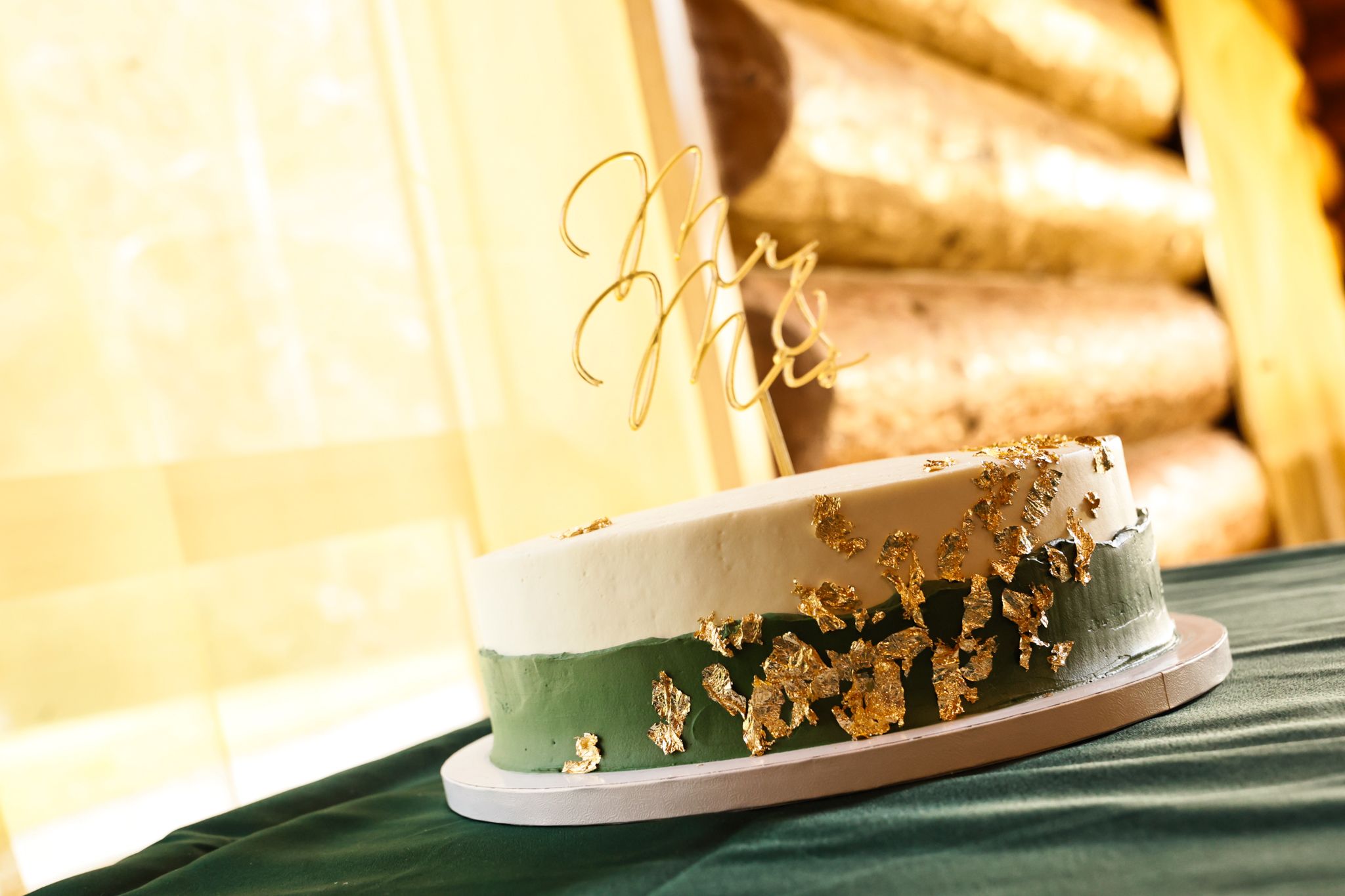 Elegant wedding cake with green and gold leaf details, Mr & Mrs topper, displayed on a green table.