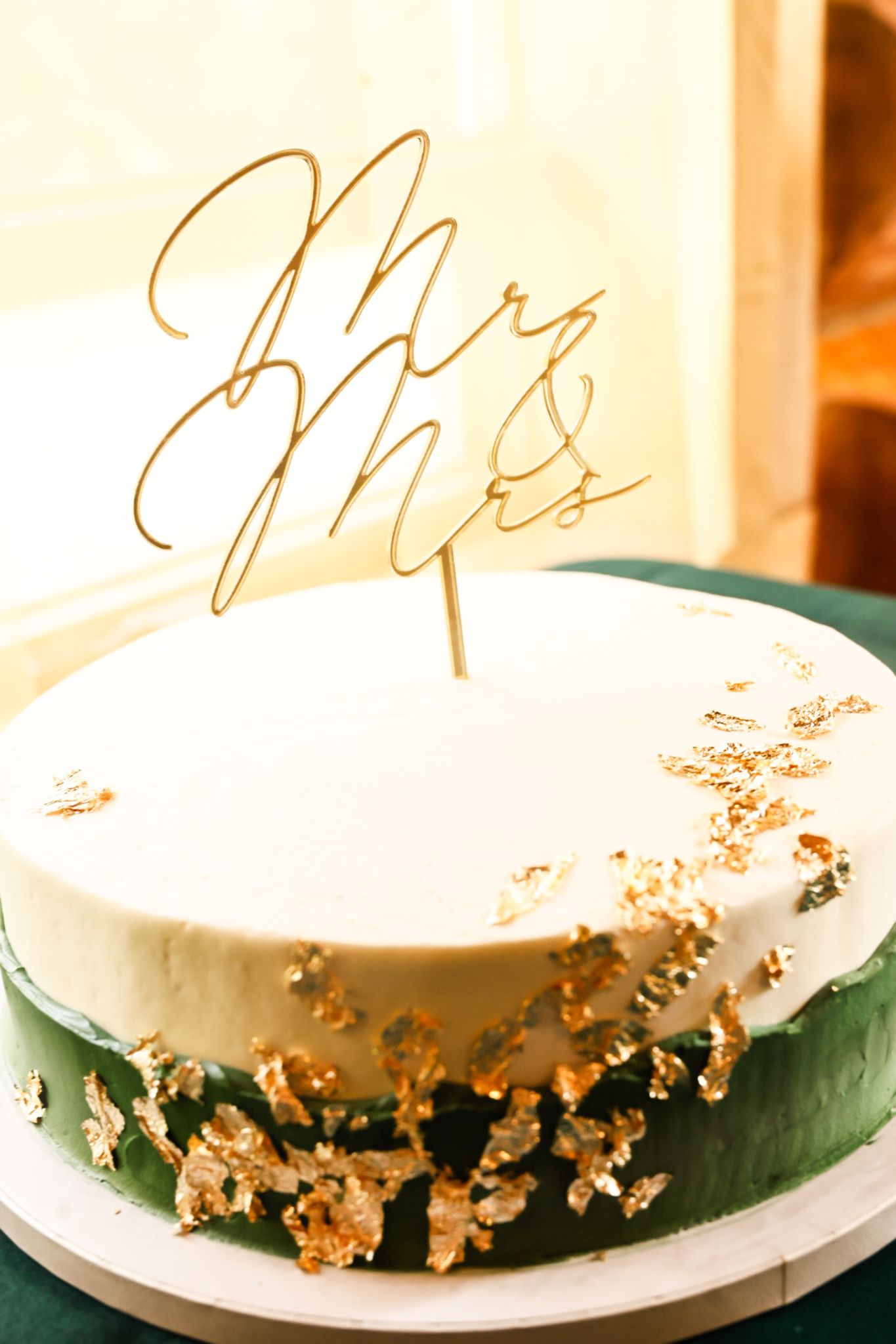 Elegant white wedding cake with gold leaf accents and a Mr. & Mrs. cake topper on a green base.