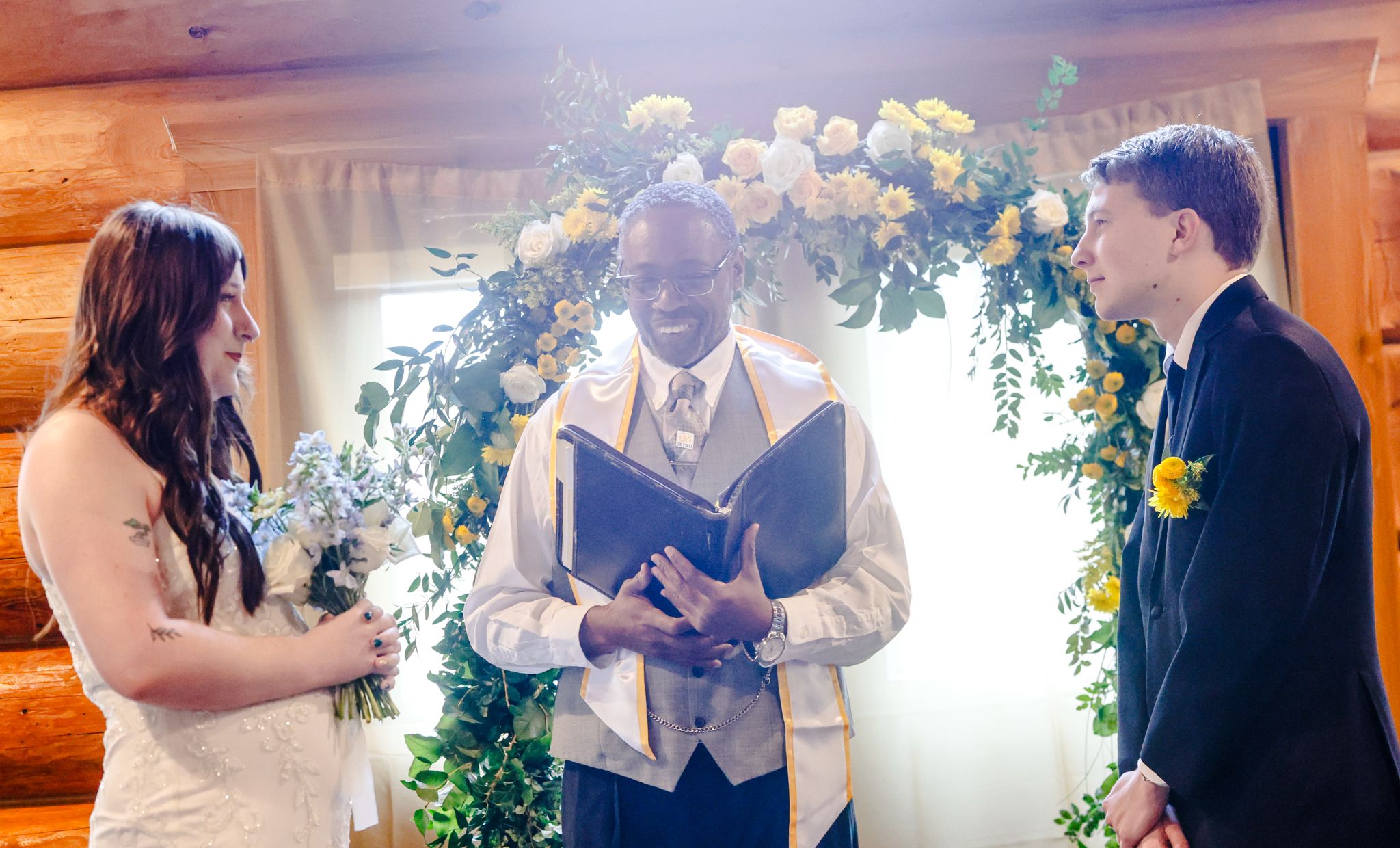 Smiling bride and groom exchange vows with officiant at rustic log cabin wedding ceremony with floral arch backdrop.
