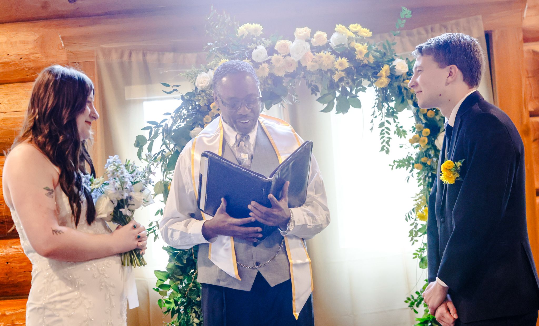 Officiant reads vows at rustic log cabin wedding ceremony with bride and groom beneath floral arch.