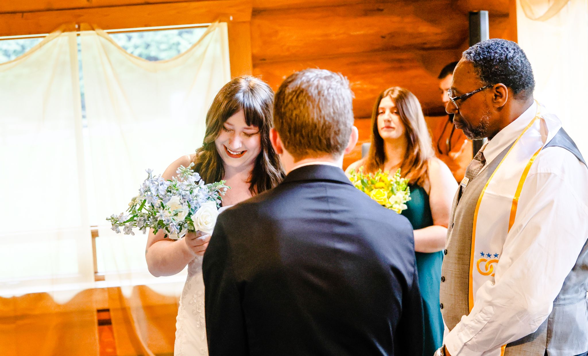 Bride holding bouquet exchanging vows with groom at rustic wedding ceremony with officiant and bridesmaid.