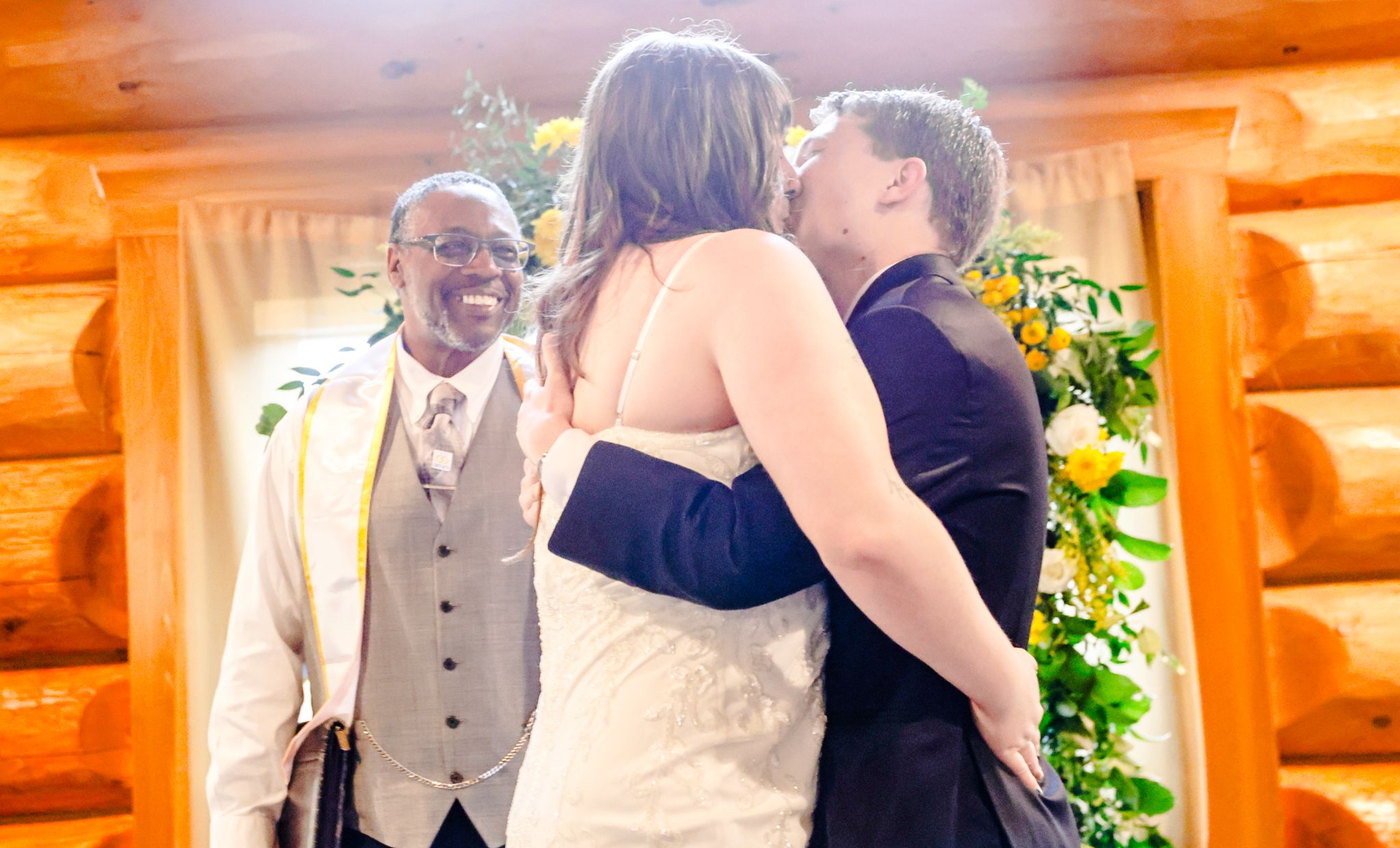 Couple sharing a kiss at wedding altar with smiling officiant in rustic log cabin venue decorated with flowers.