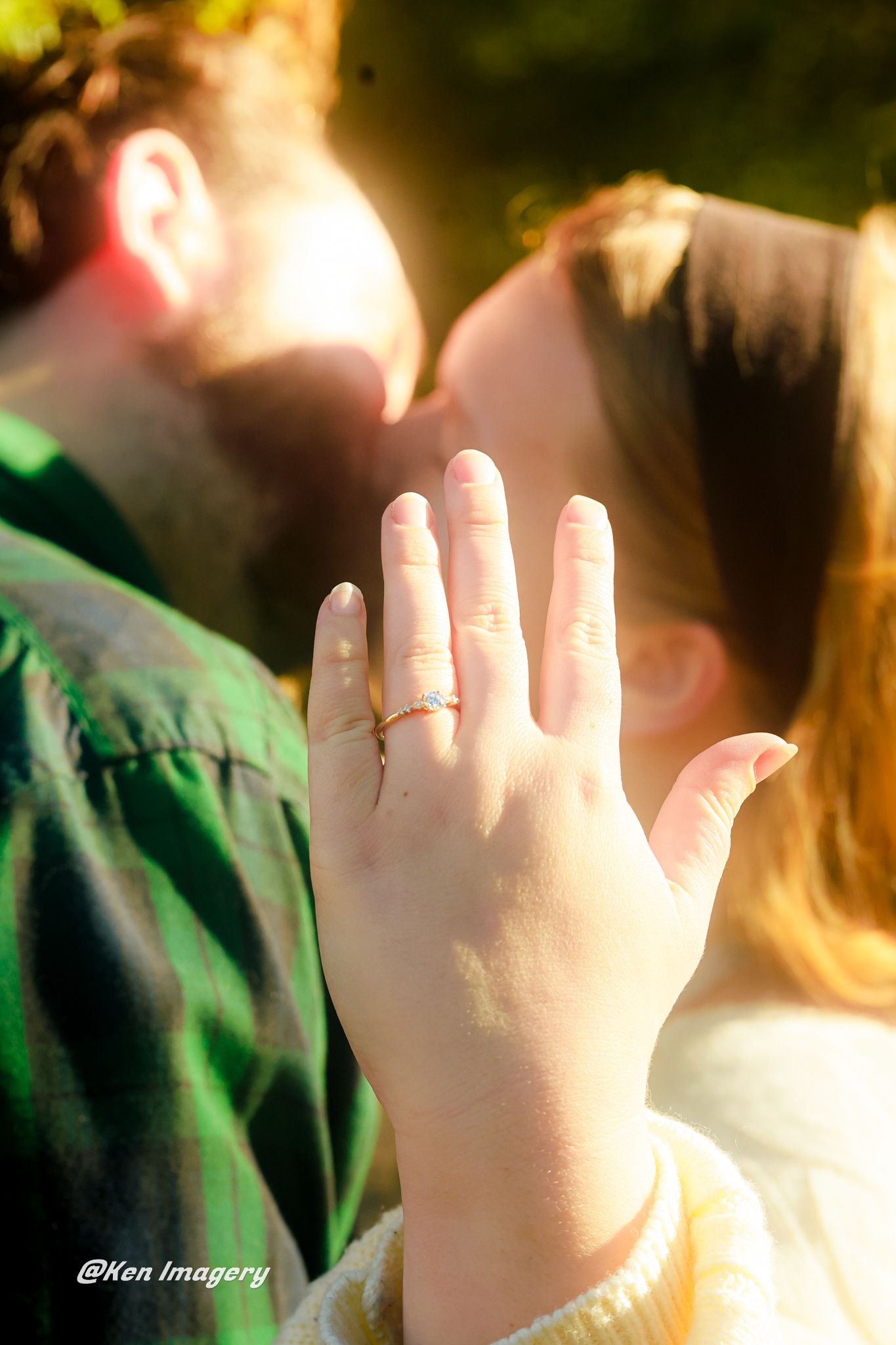 Couple kissing in golden sunlight while woman displays engagement ring with diamond band toward camera.