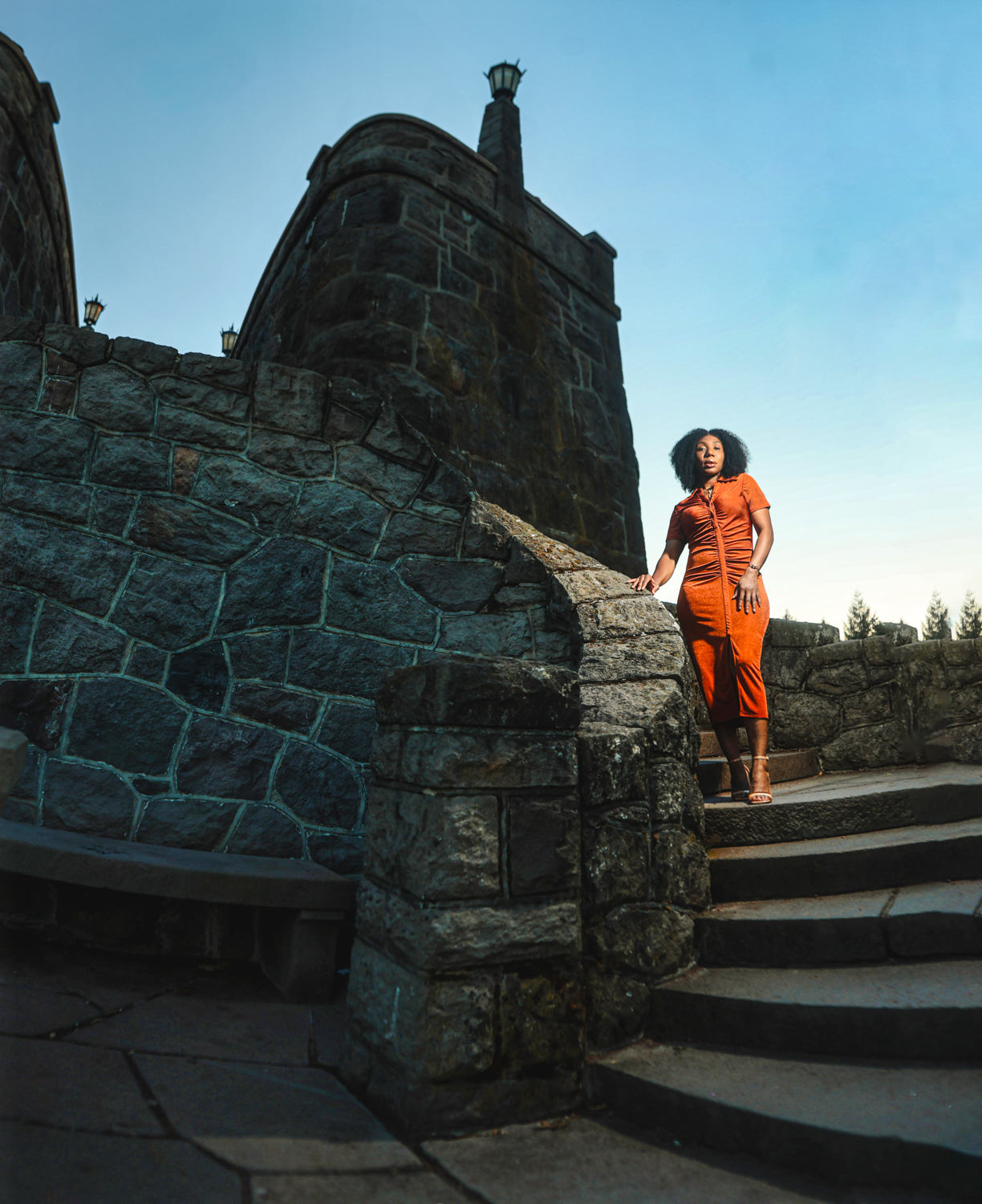 A woman in an orange outfit stands on stone steps of a historic castle at dusk.