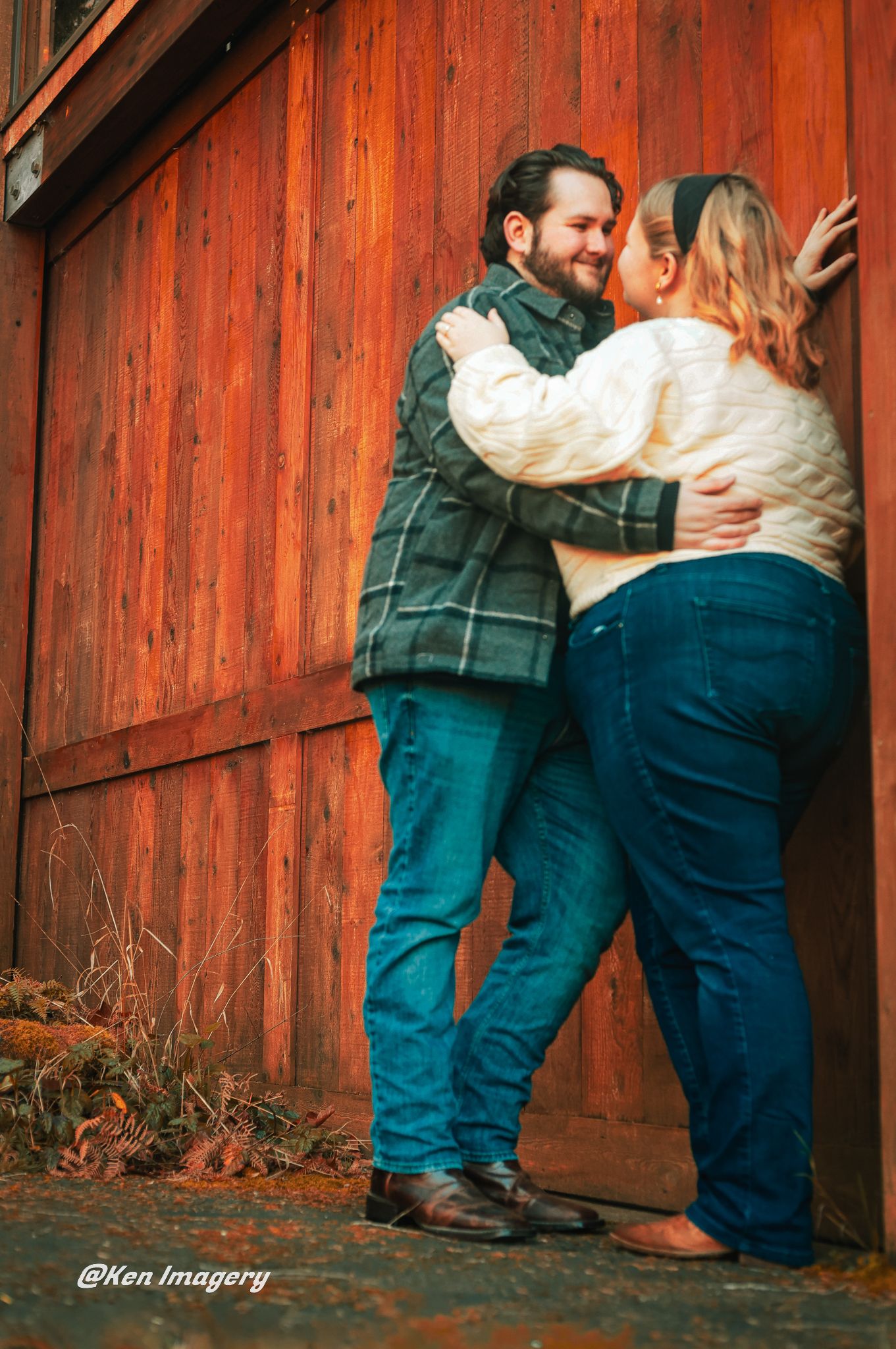 Couple embracing and smiling against a rustic red barn wall, wearing jeans and casual sweaters outdoors.
