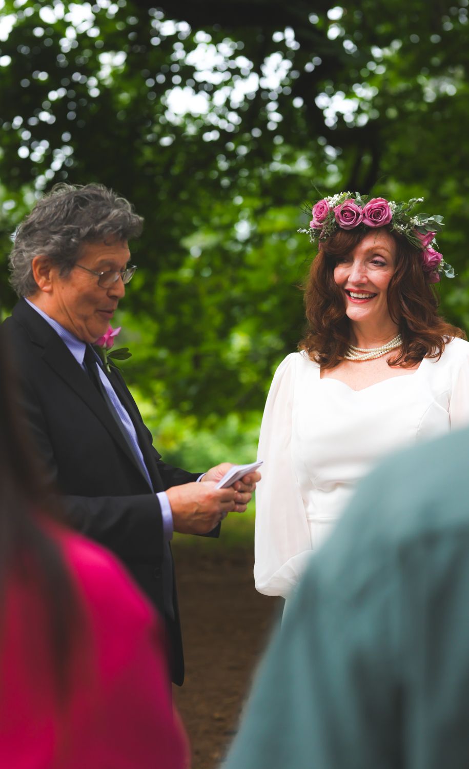 Smiling bride in white dress and pink flower crown exchanges vows with officiant outdoors at garden wedding ceremony.