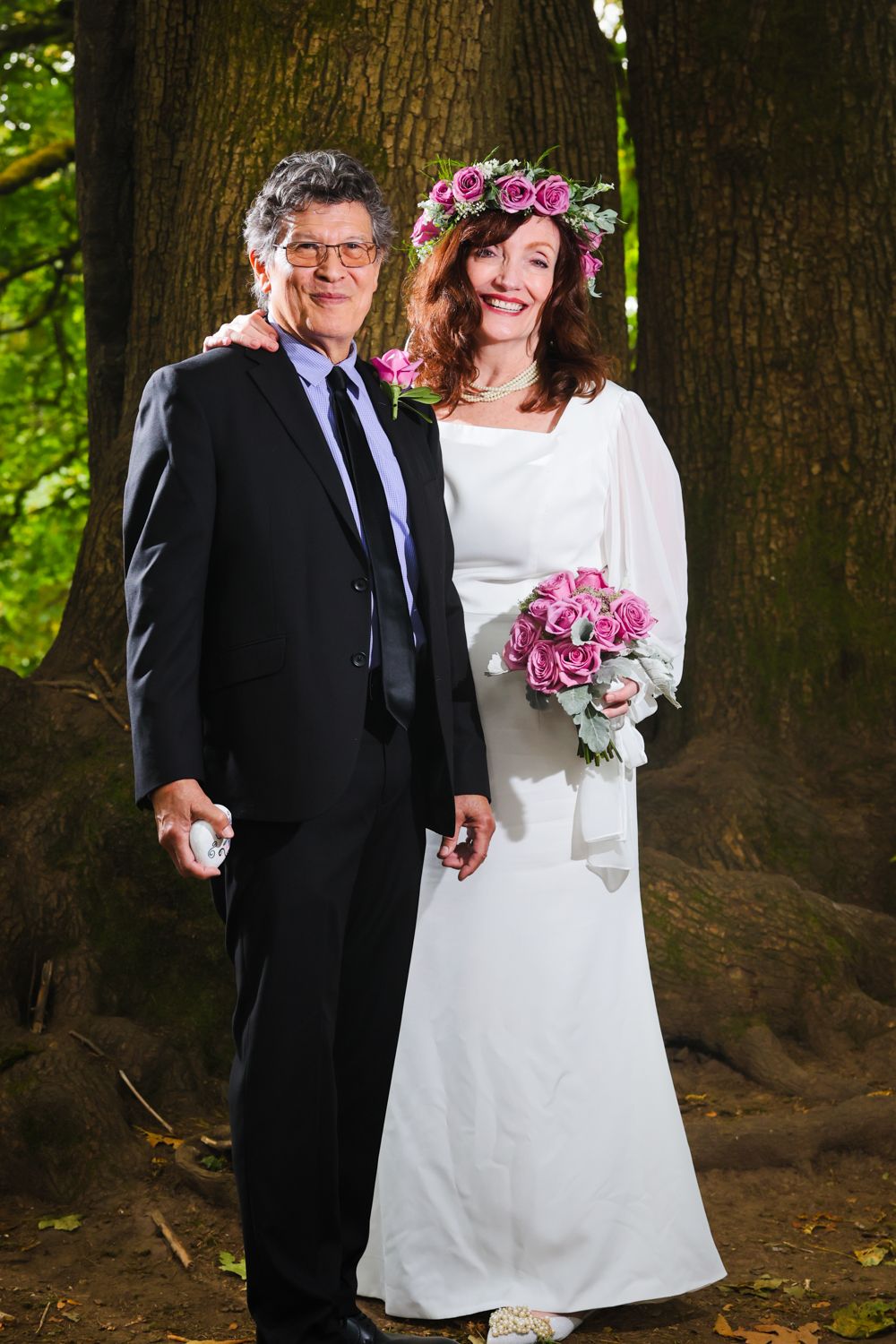 Bride in white gown with pink flower crown holds bouquet beside groom in black suit among trees.