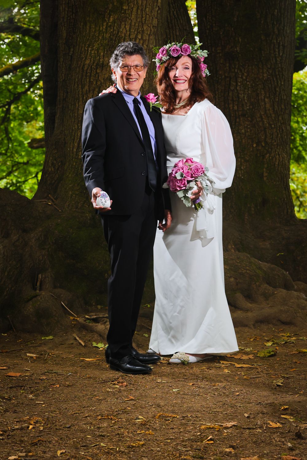 Mature couple in wedding attire pose outdoors among trees, bride in white gown with pink floral crown holding bouquet.