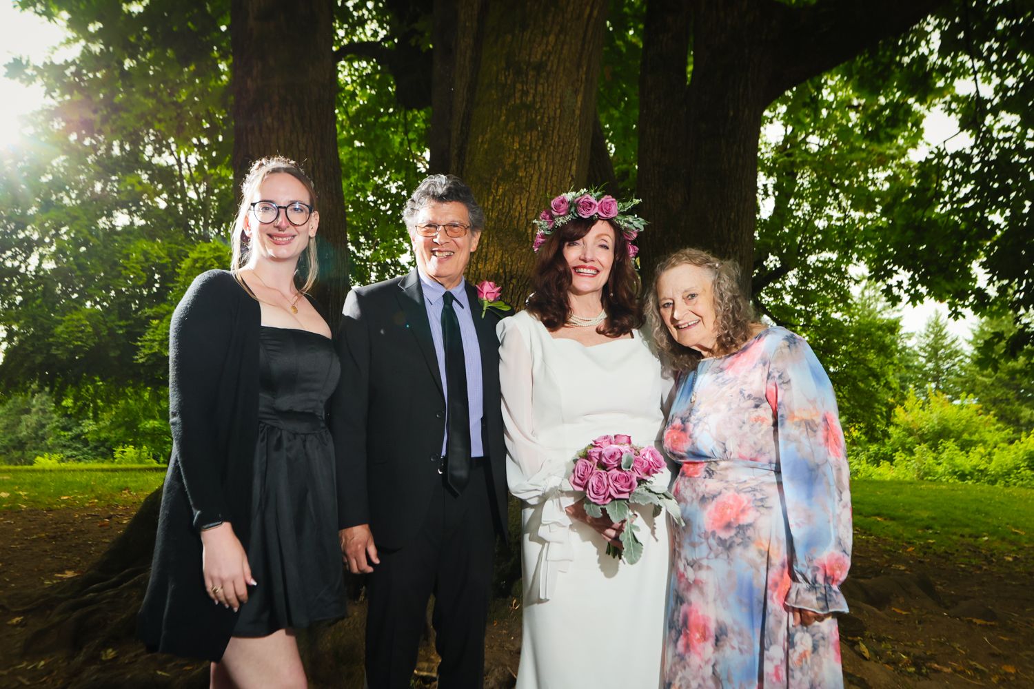 Wedding party poses outdoors near large tree, bride in white dress with floral crown holding bouquet with family.