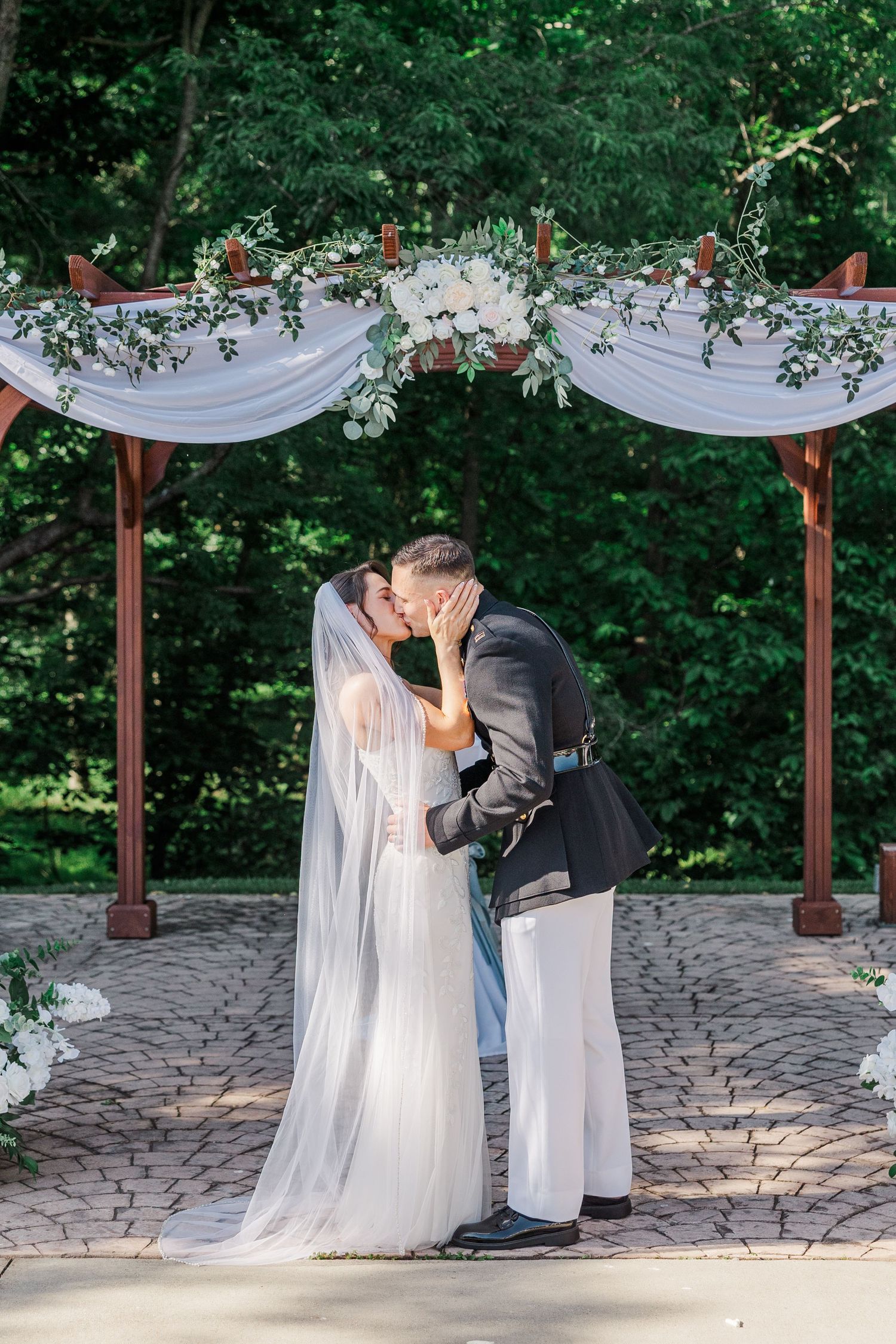 Bride and groom share first kiss under floral arch at outdoor wedding ceremony surrounded by lush green trees.