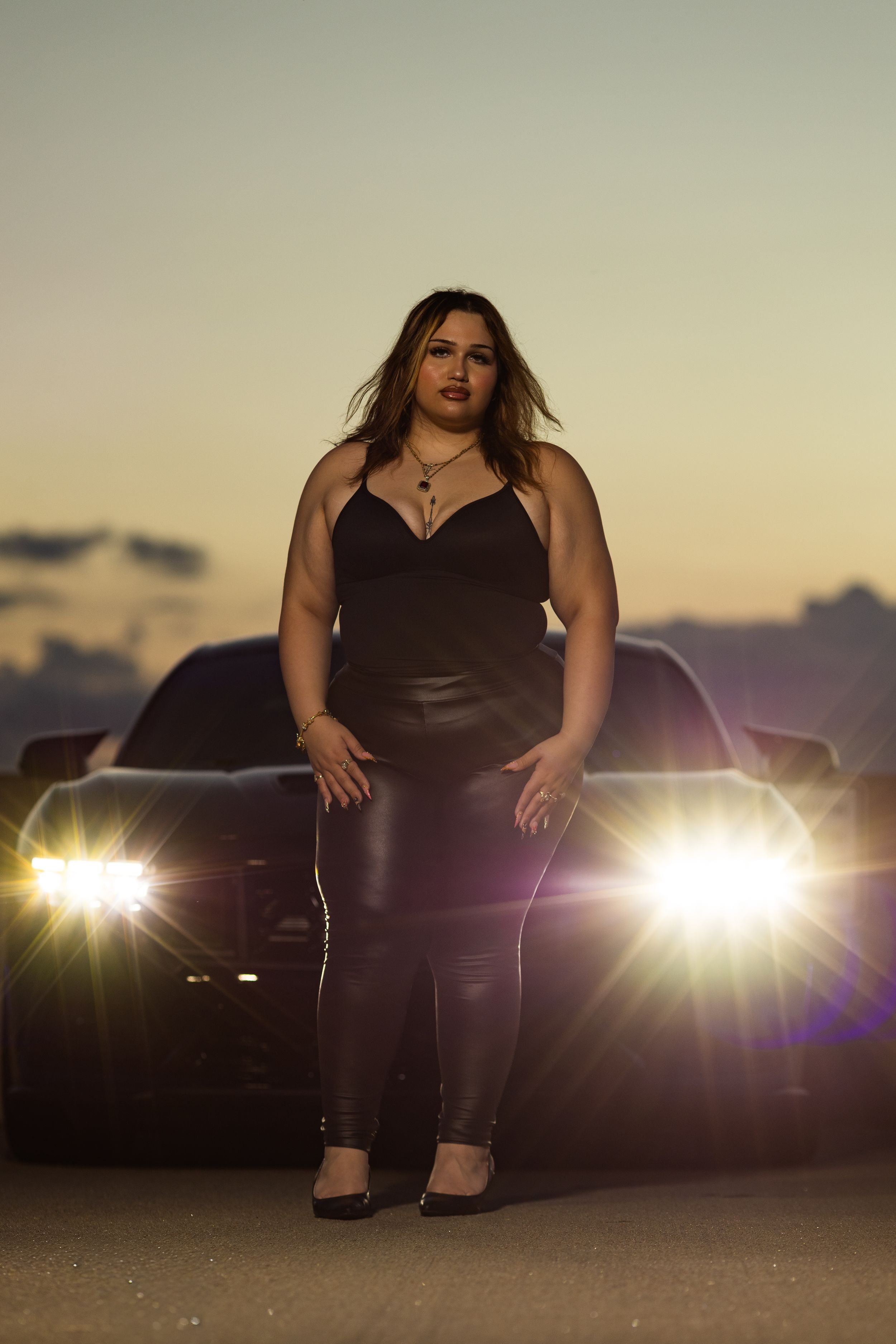 Woman in black outfit posing confidently in front of a car with bright headlights at sunset.