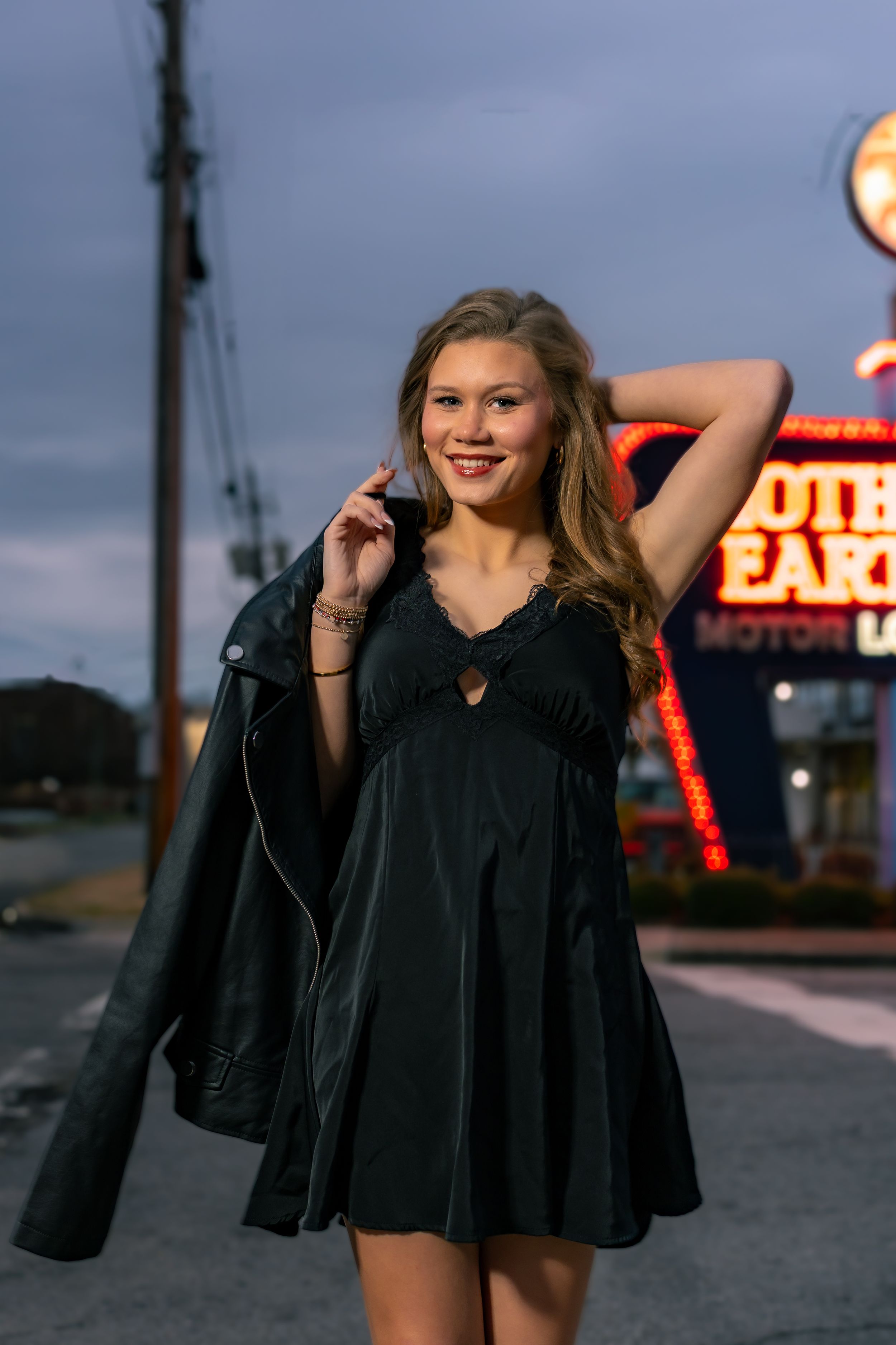 Young blonde woman in black dress and leather jacket poses smiling before a neon motel sign at dusk.
