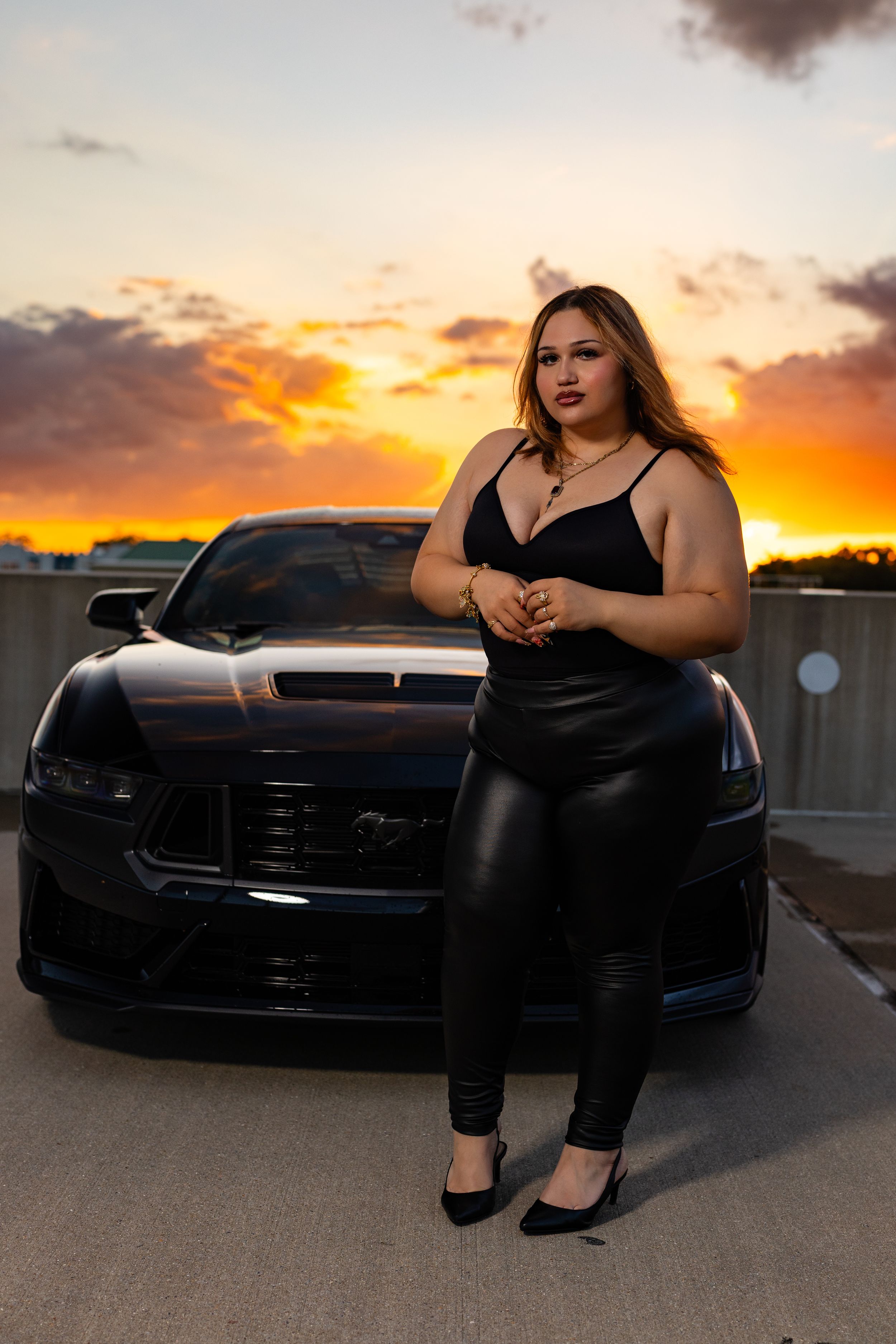 Woman in black outfit poses against a dark Ford Mustang at sunset with vibrant orange sky.