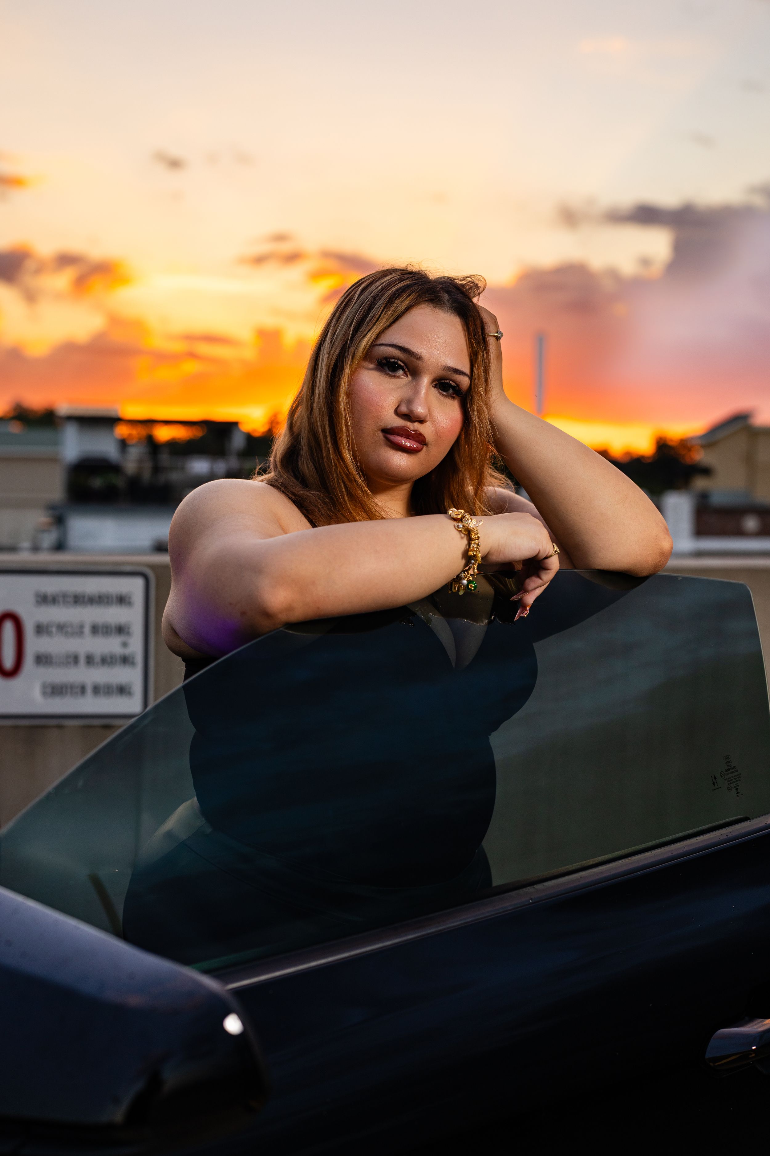 Woman leaning on car door at golden hour sunset, wearing gold bracelet, posing on rooftop parking lot.