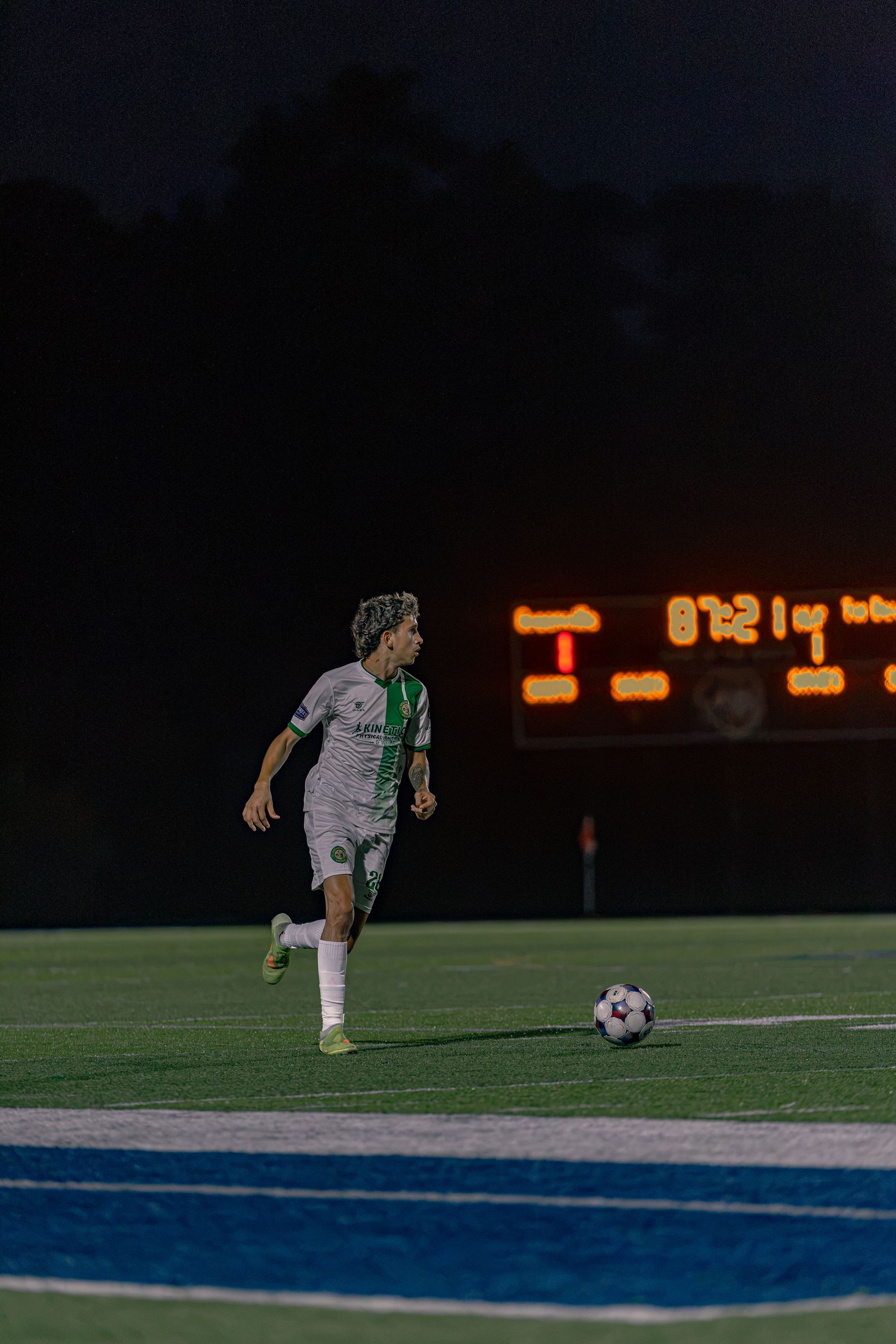 Soccer player in white uniform dribbling ball on field at night with illuminated scoreboard in background.