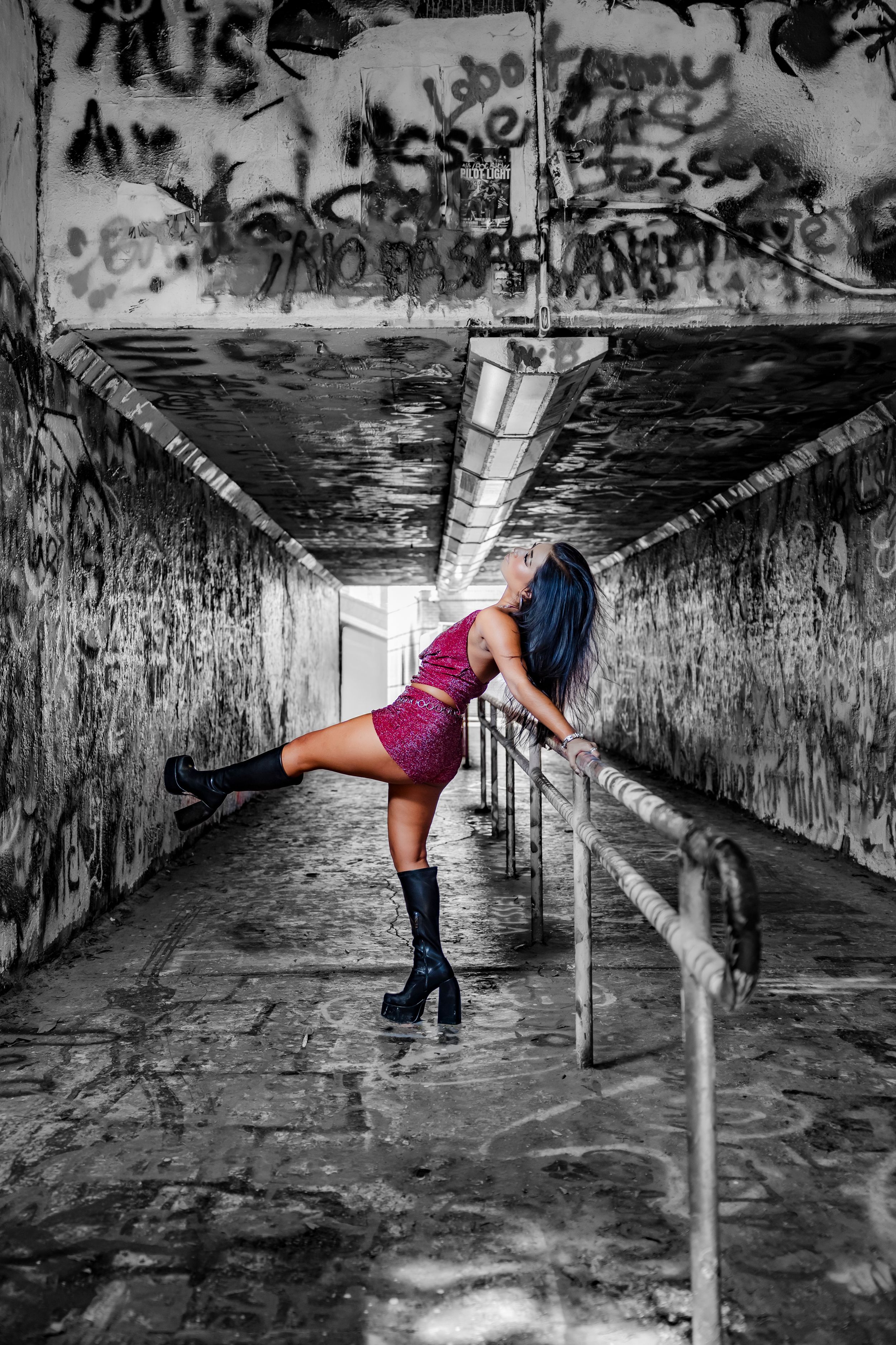 Woman in pink dress and black boots poses dramatically on railing in graffiti-covered tunnel with selective color effect.