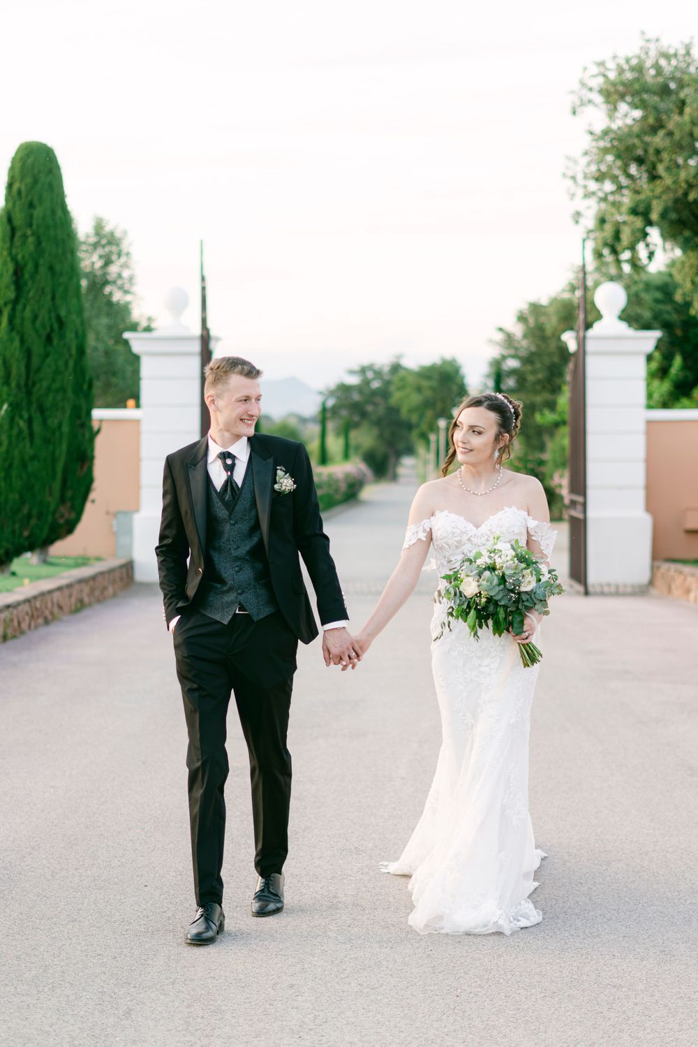 Bride and groom share a romantic kiss among lush vineyard rows at golden hour, bride holding white floral bouquet.