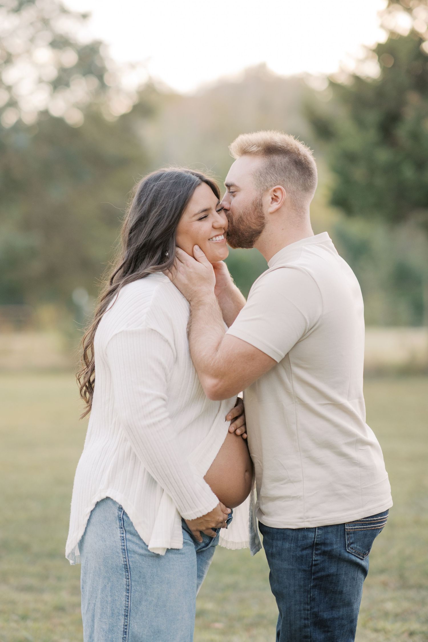 A couple shares a tender moment outdoors, man kissing woman's cheek while cradling her face in a sunny field.
