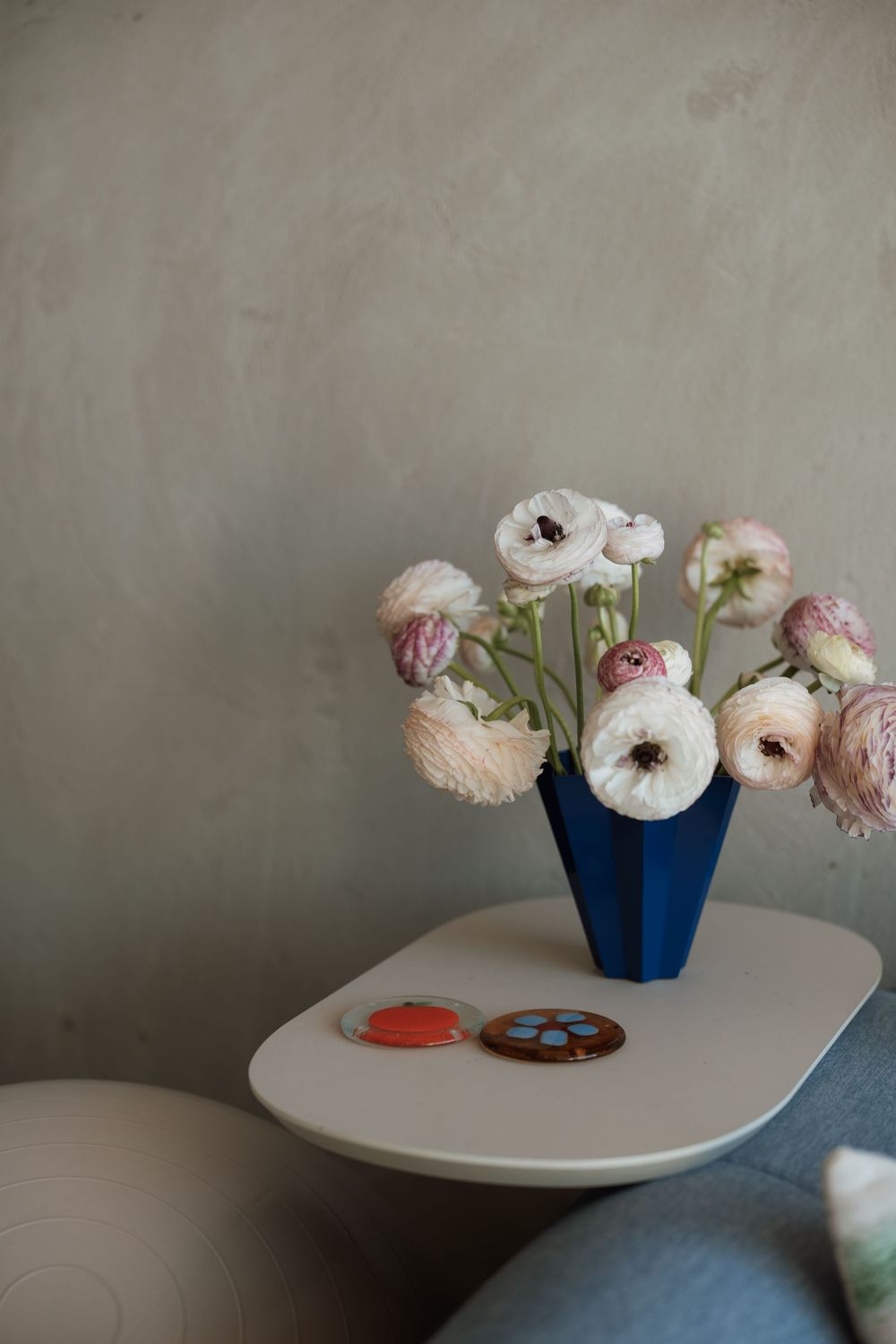 Blue geometric vase with white and pink ranunculus flowers on a modern side table with decorative coasters.