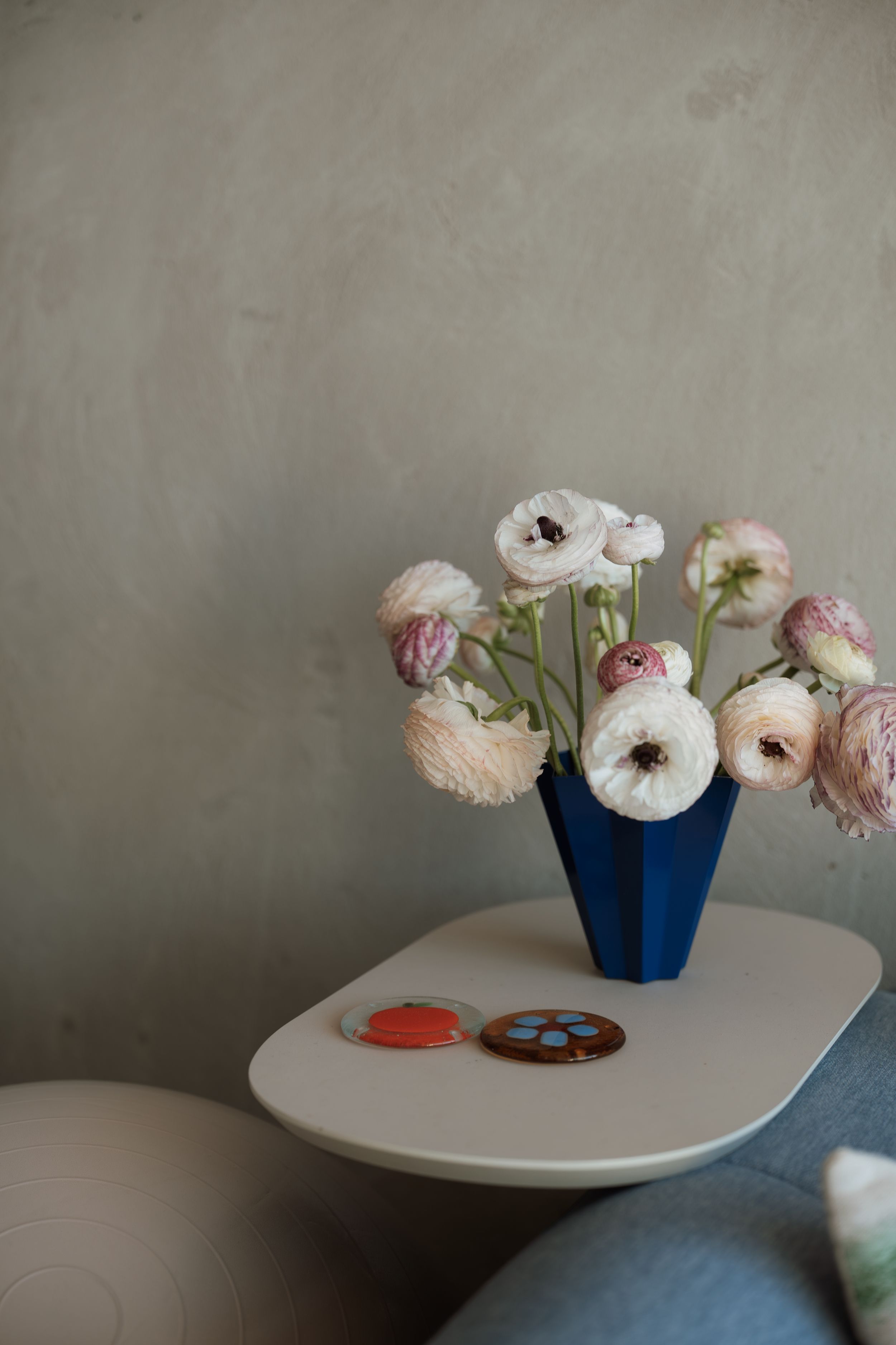 Blue geometric vase with white and pink ranunculus flowers on a modern side table with decorative coasters.