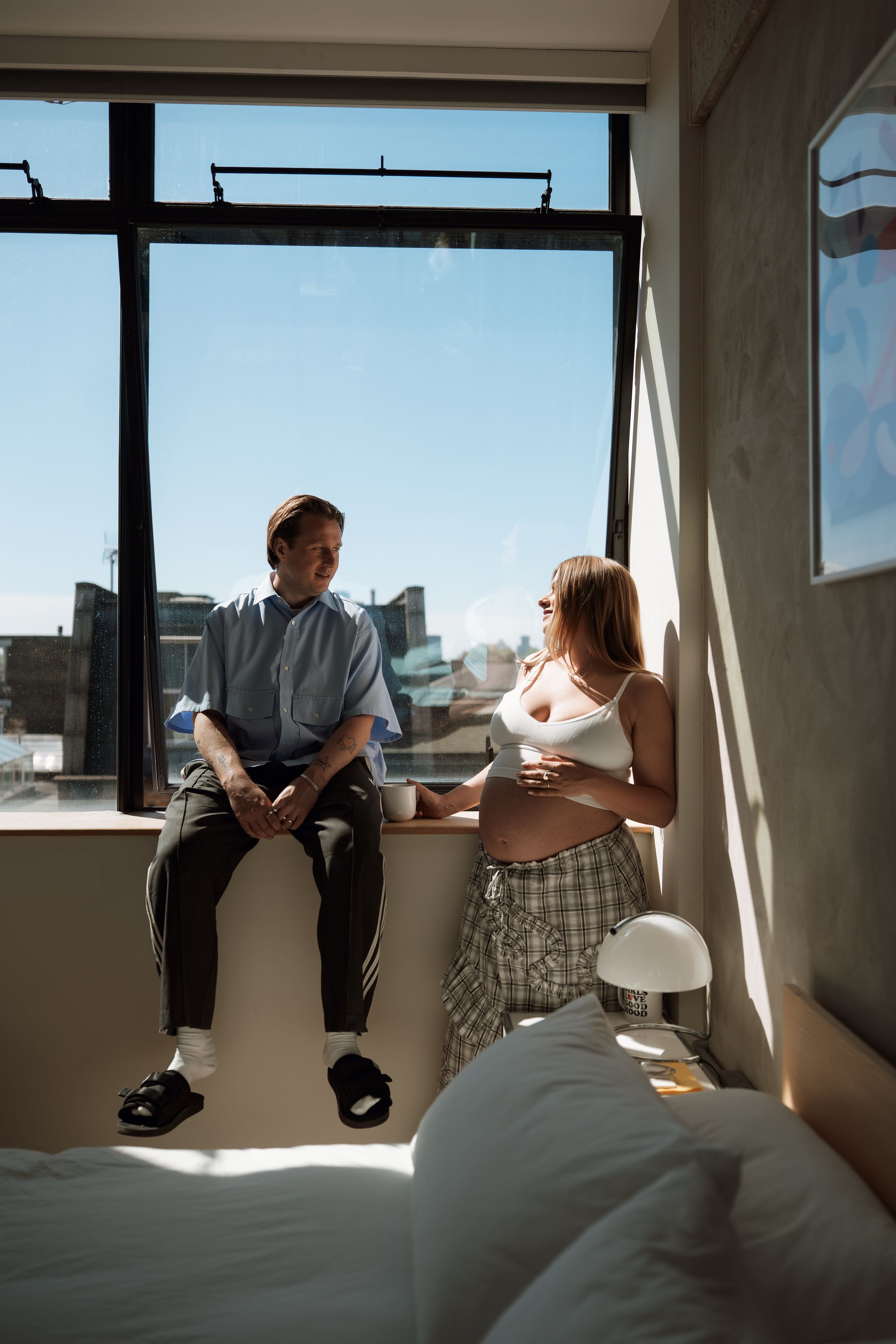 A couple shares a tender moment by a sunny window, the woman visibly pregnant, in a modern bedroom with city views.