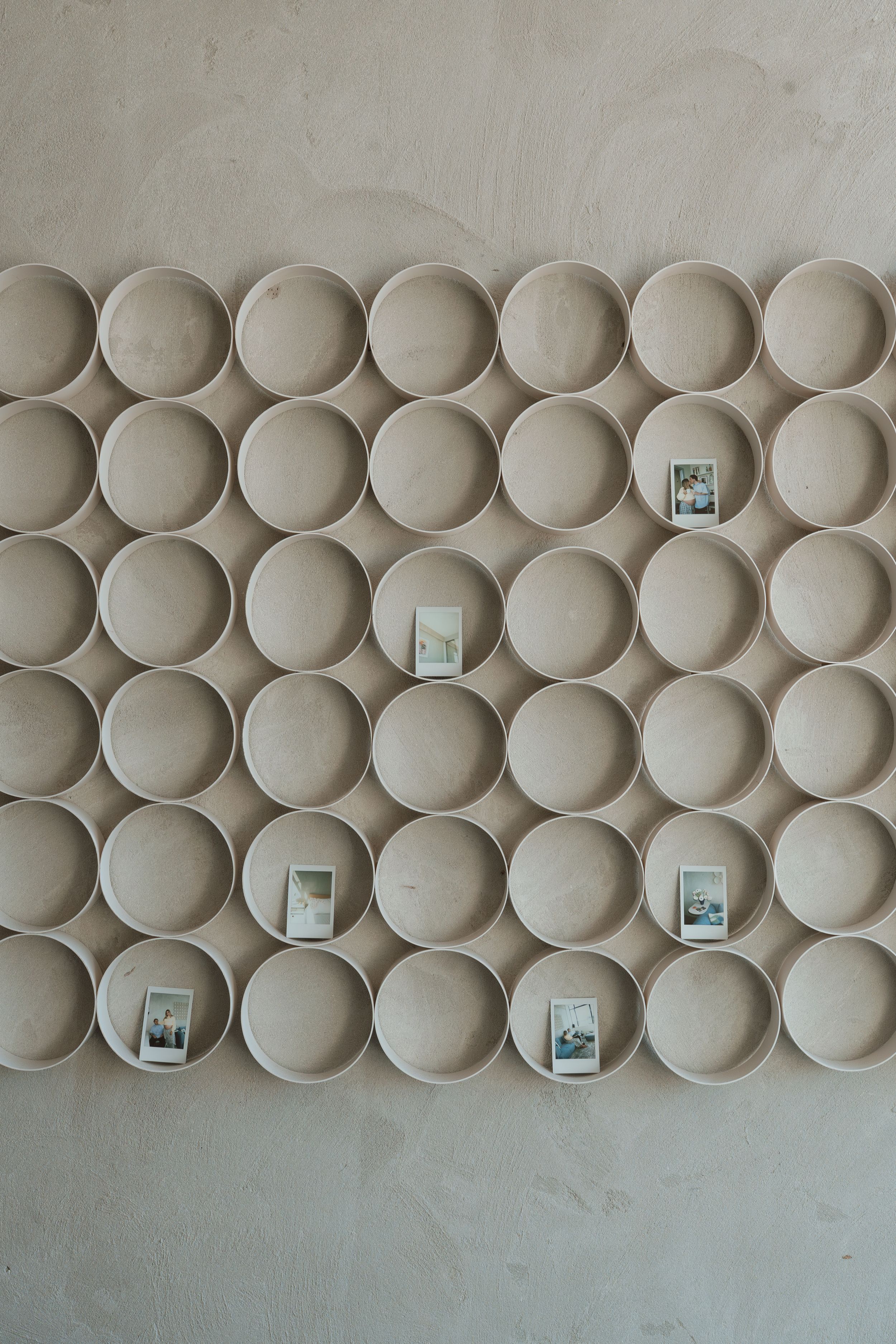 Wall display of circular wooden rings arranged in a grid pattern with Polaroid photos placed in select compartments.