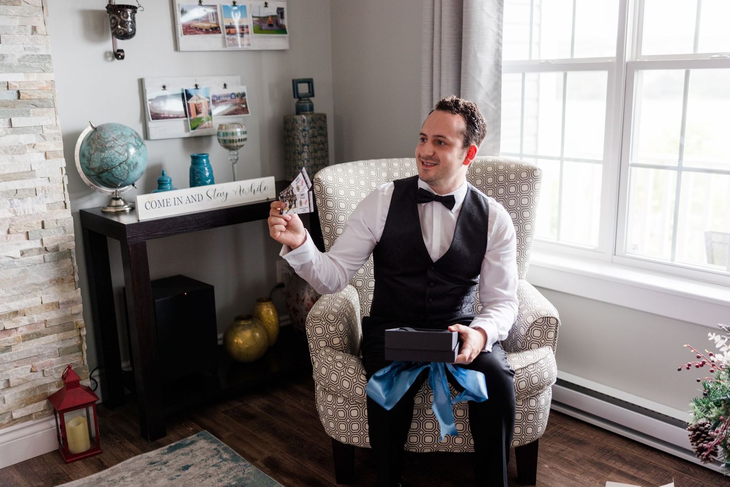 A man in a tuxedo vest and bow tie sits in a patterned armchair, holding a card in a bright living room.