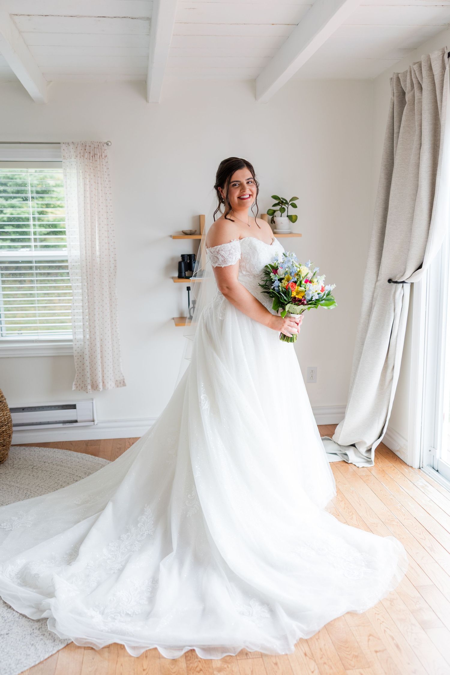 Bride in off-shoulder white ball gown with long train holds colorful bouquet in bright white room.