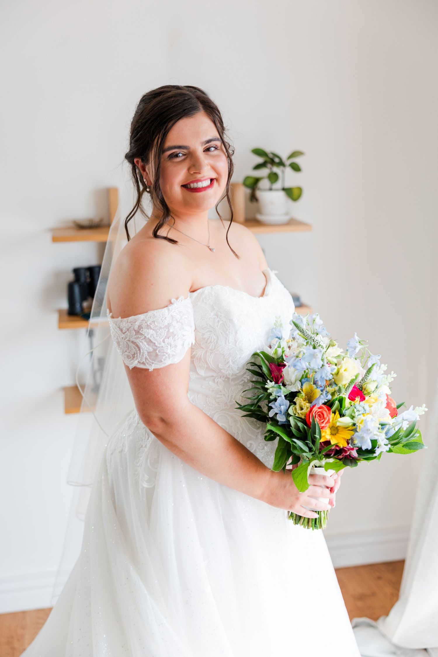 Smiling bride in off-shoulder lace wedding dress holding colorful bouquet with blue, yellow, and pink flowers indoors.