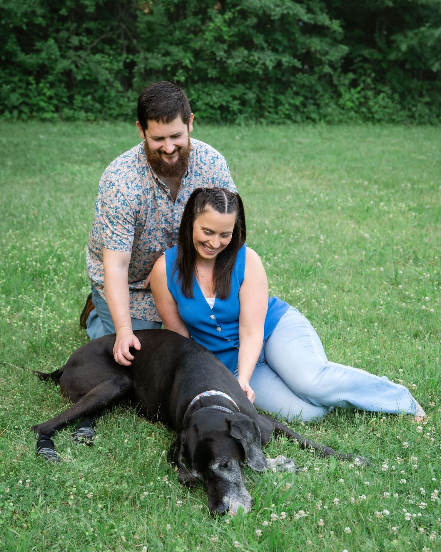 Couple smiling and sitting on green grass with a large black Great Dane dog outdoors near trees.