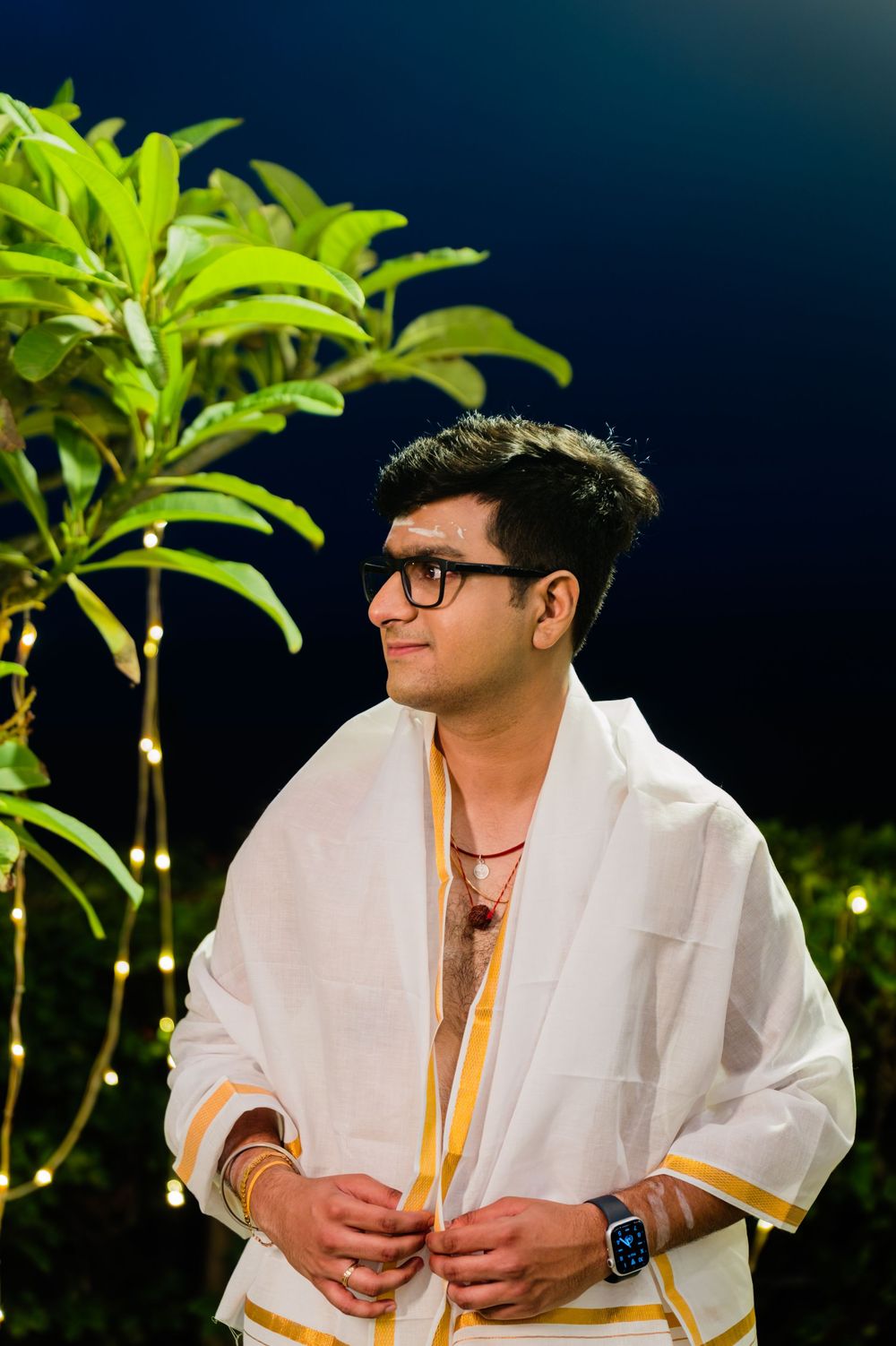 Young Indian man in traditional white dhoti with gold border and glasses at an outdoor evening ceremony.