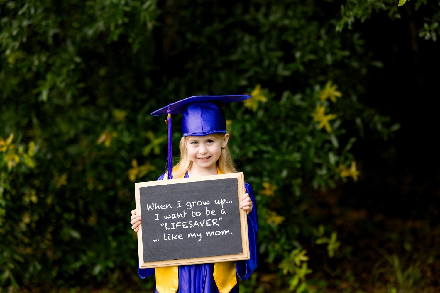 Young girl in blue graduation cap and gown holds chalkboard sign reading she wants to be a lifesaver like her mom.
